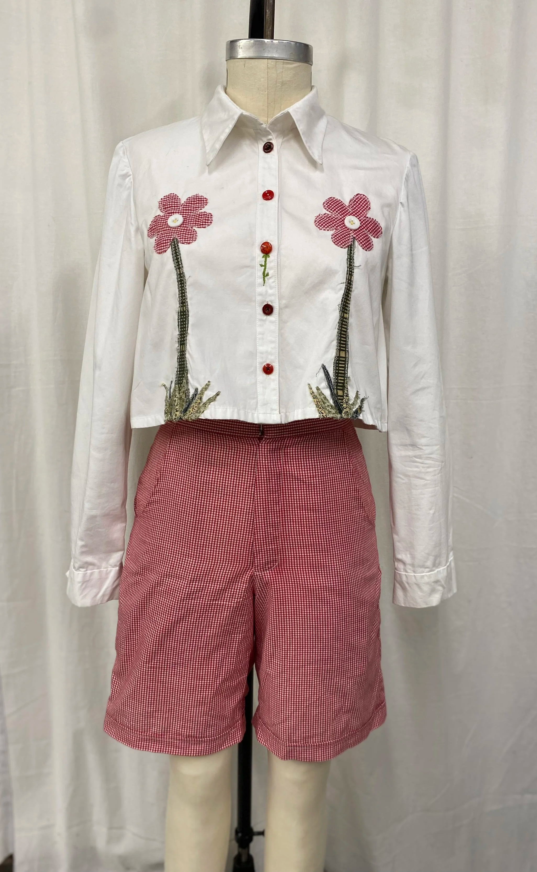 A mannequin dressed in a white button-up shirt with red buttons and embroidery of pink flowers with green stems, paired with red and white checkered shorts.