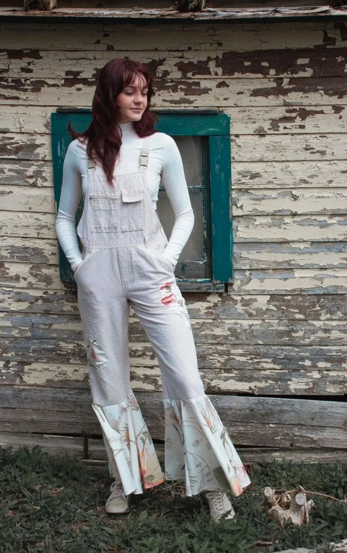 A young woman with long dark hair and light skin stands in front of a weathered, peeling wooden wall with a small window behind her. She is wearing a white long-sleeve top and light-colored overalls with distressed details and a floral pattern at the flared pant bottoms. She has her hands in her pockets and is looking to her left.