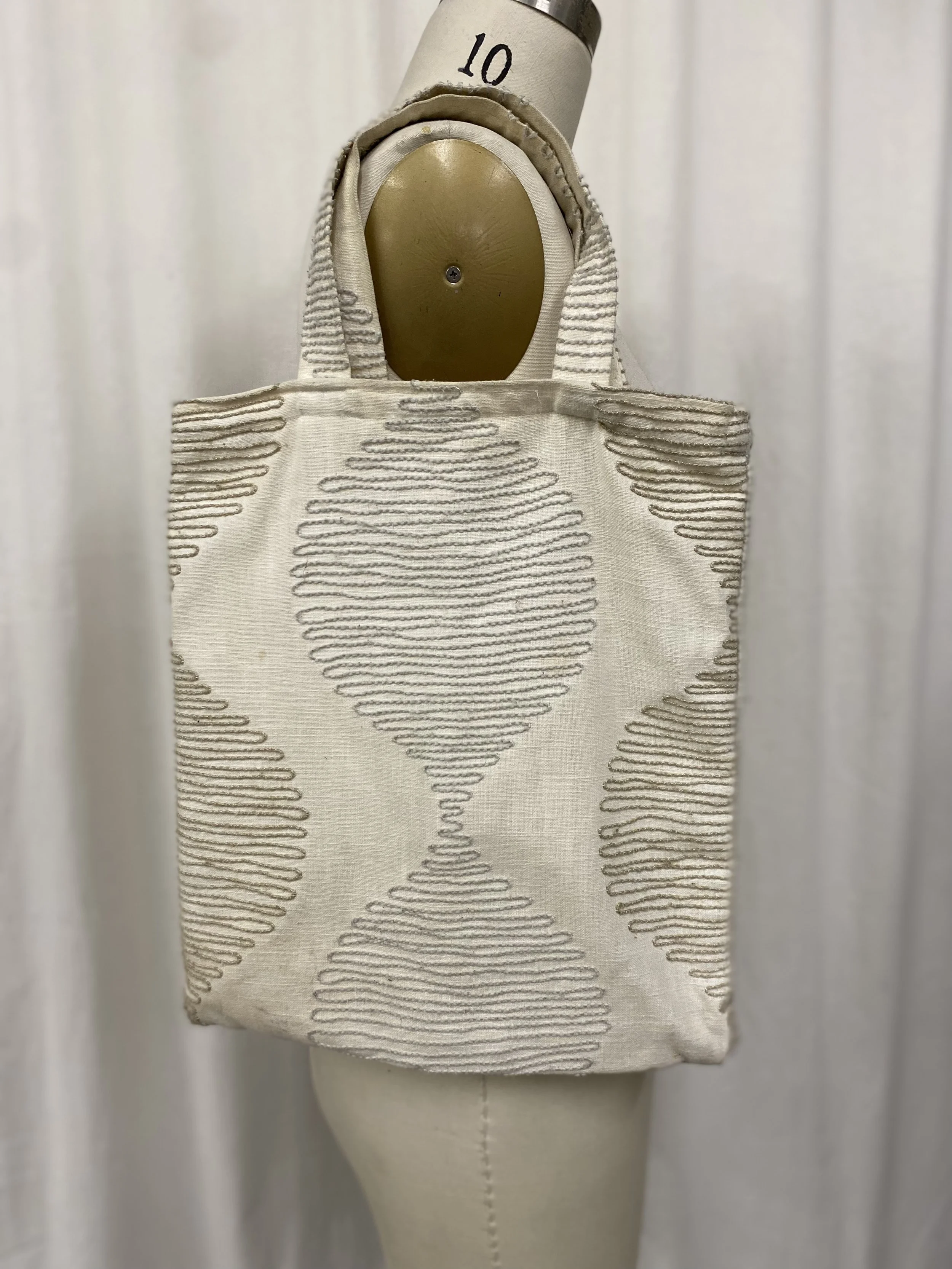 A cream-colored tote bag with wavy stitched patterns hanging on a dress form, set against a plain white background.
