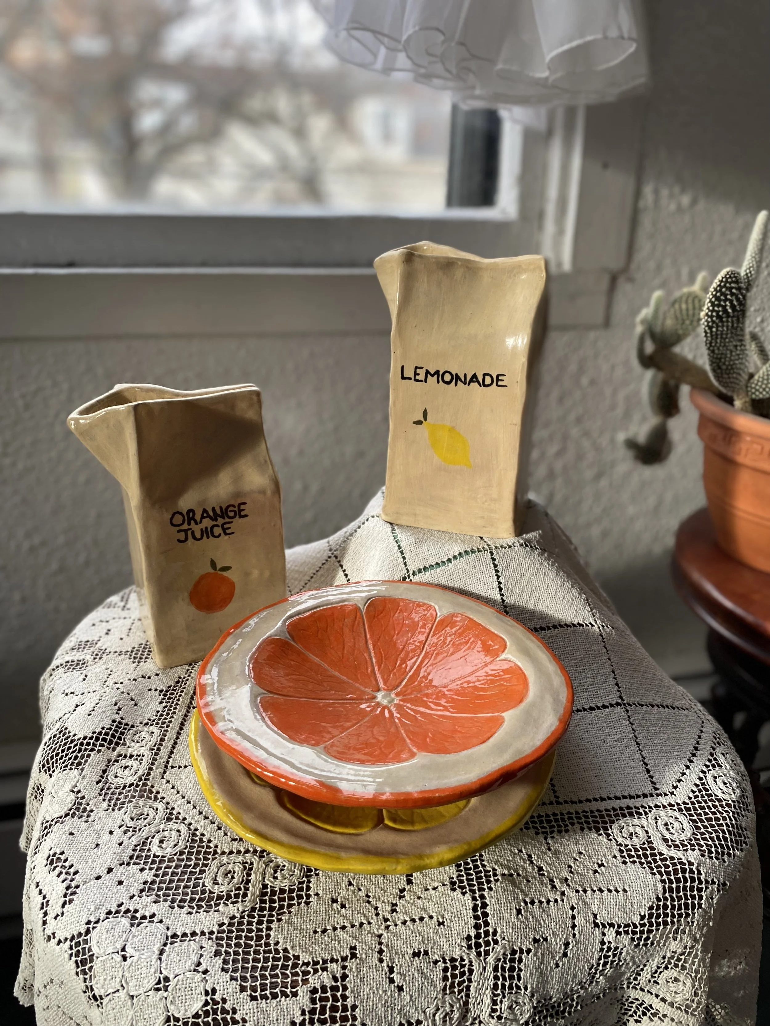 Decorative ceramic plates resembling orange and lemon slices, placed on a lace tablecloth near a window with two small ceramic pitchers labeled 'Orange Juice' and 'Lemonade'.
