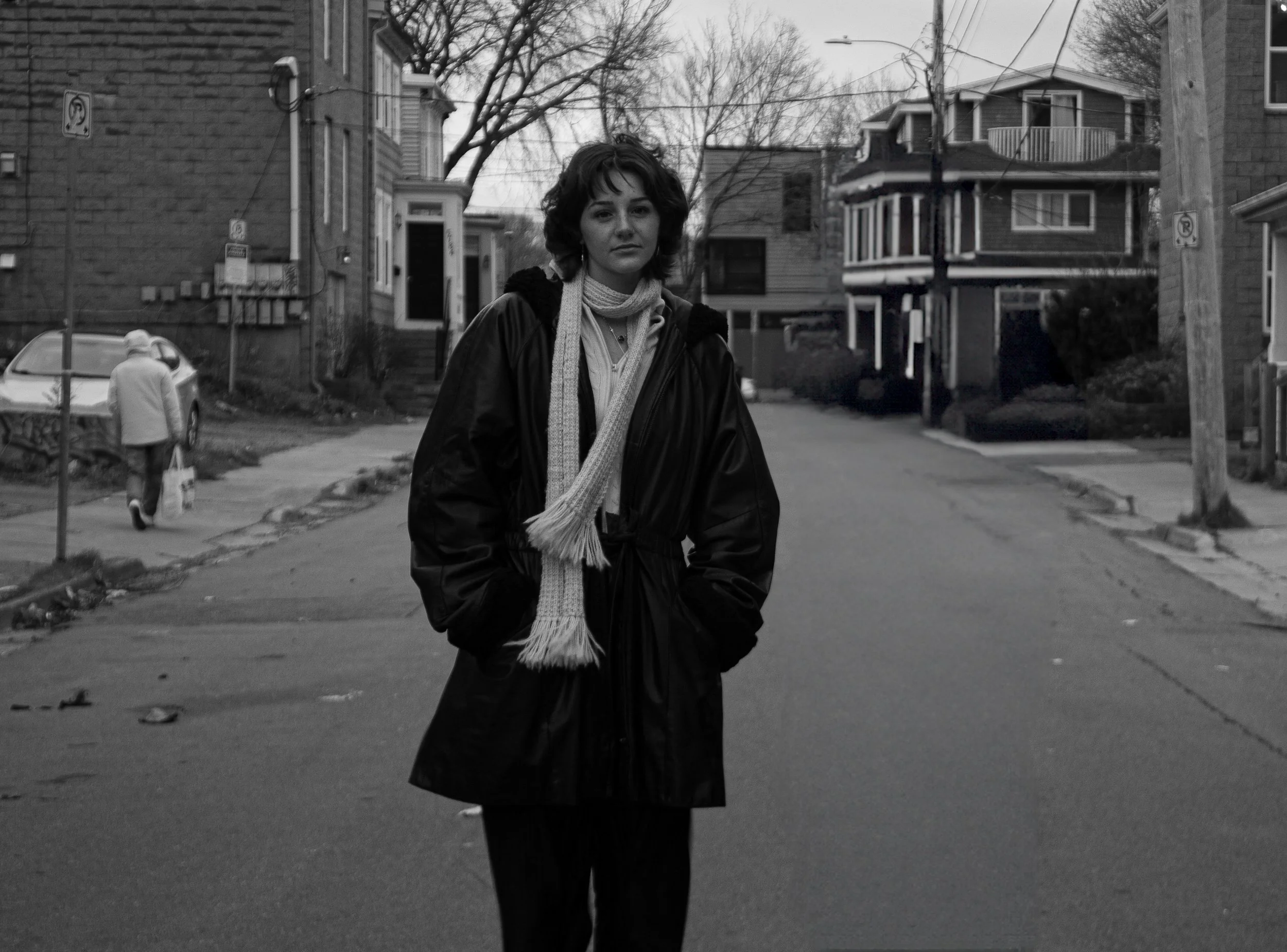 A woman with curly hair wearing a dark coat and a light scarf stands in the middle of a residential street in black and white. In the background, there are houses, trees, a person walking away carrying a bag, and street signs.