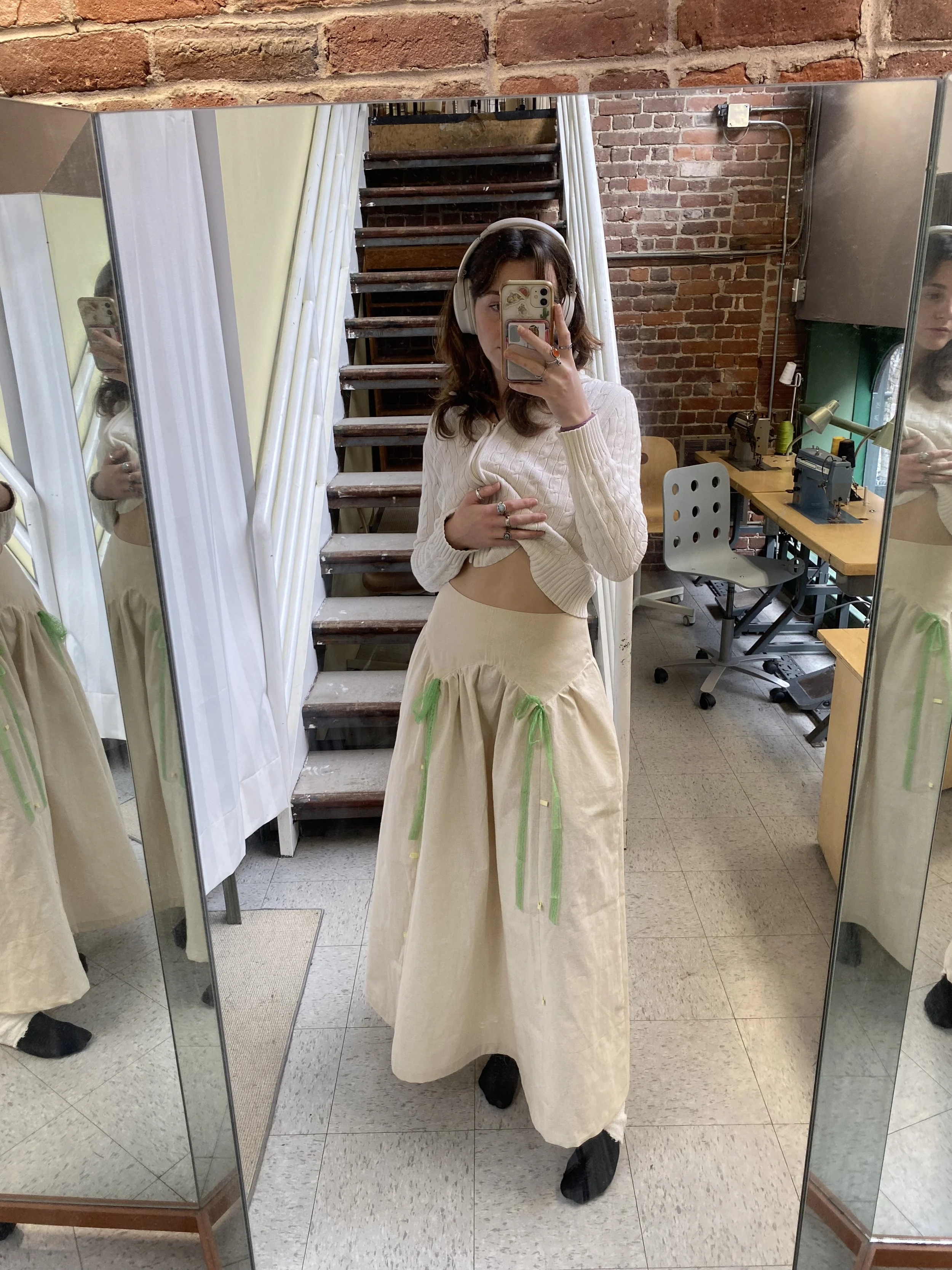 A woman taking a mirror selfie in a room with a brick wall, stairs, and sewing machines. She is wearing a cream-colored sweater, a cream-colored skirt with green ribbon accents, and black shoes, with headphones on.