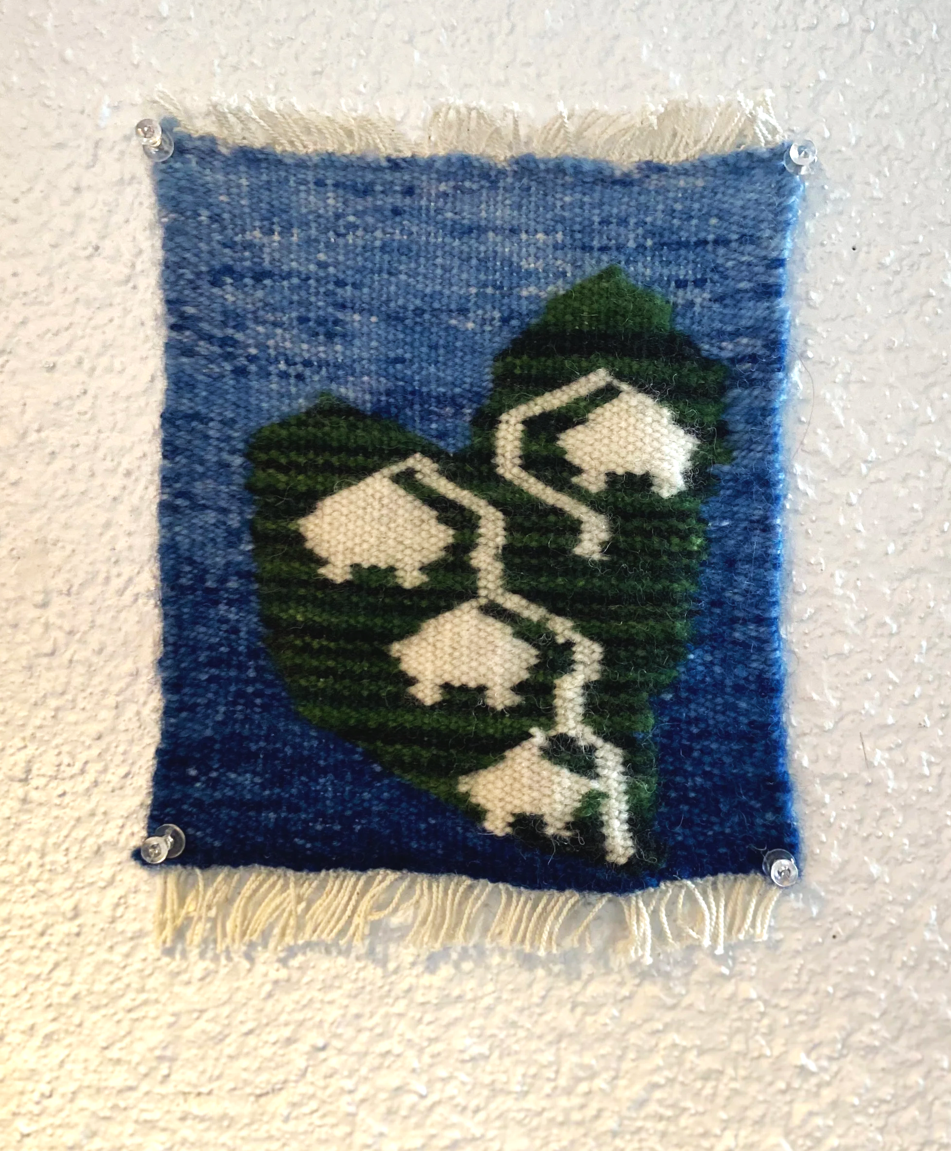 A woven textile wall hanging featuring a stylized green leaf with white geometric shapes and a black line, mounted on a textured white wall.