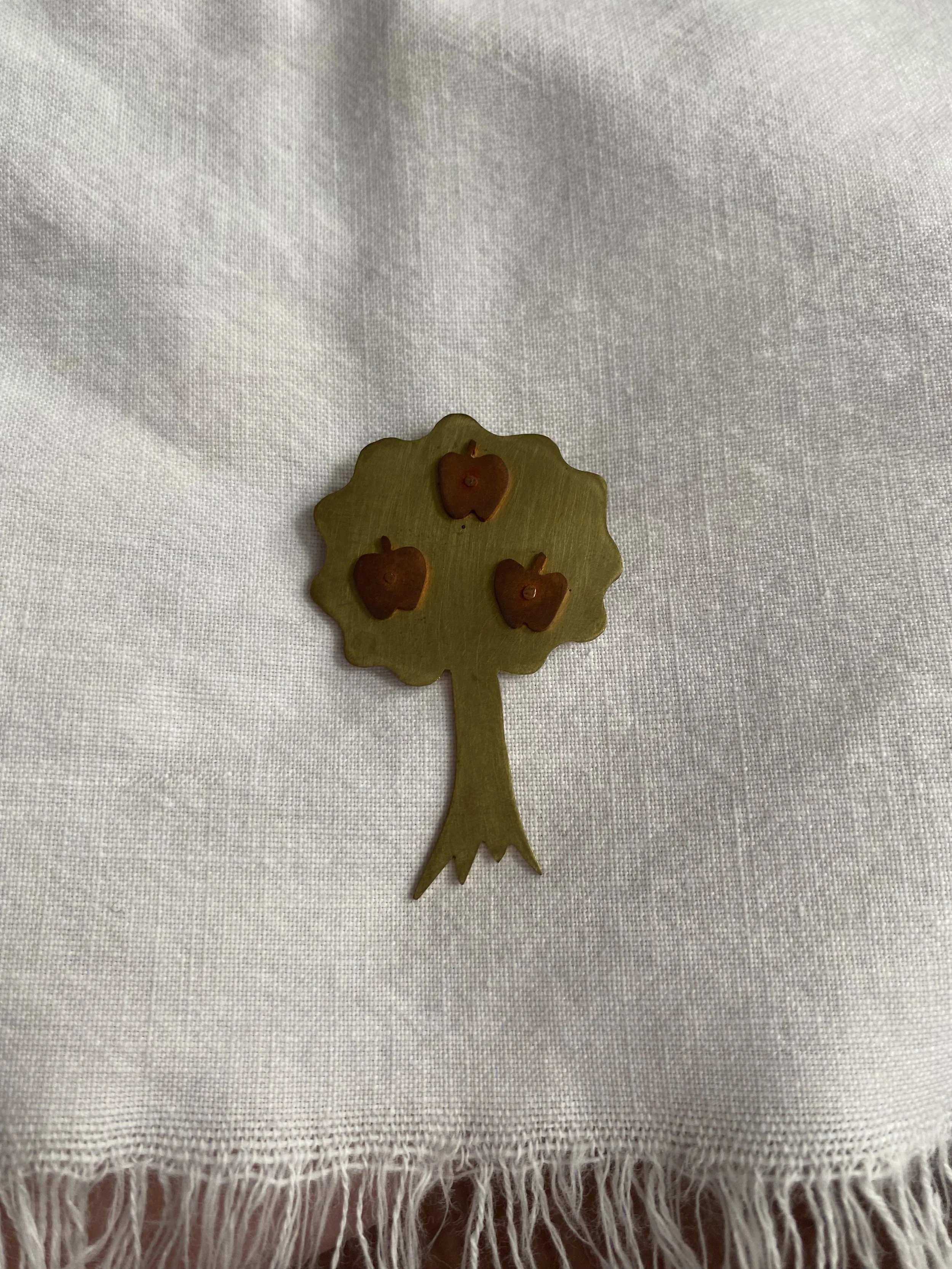 A small, decorative tree with brown apples on it, made of wood or metal, placed on a white fabric background.