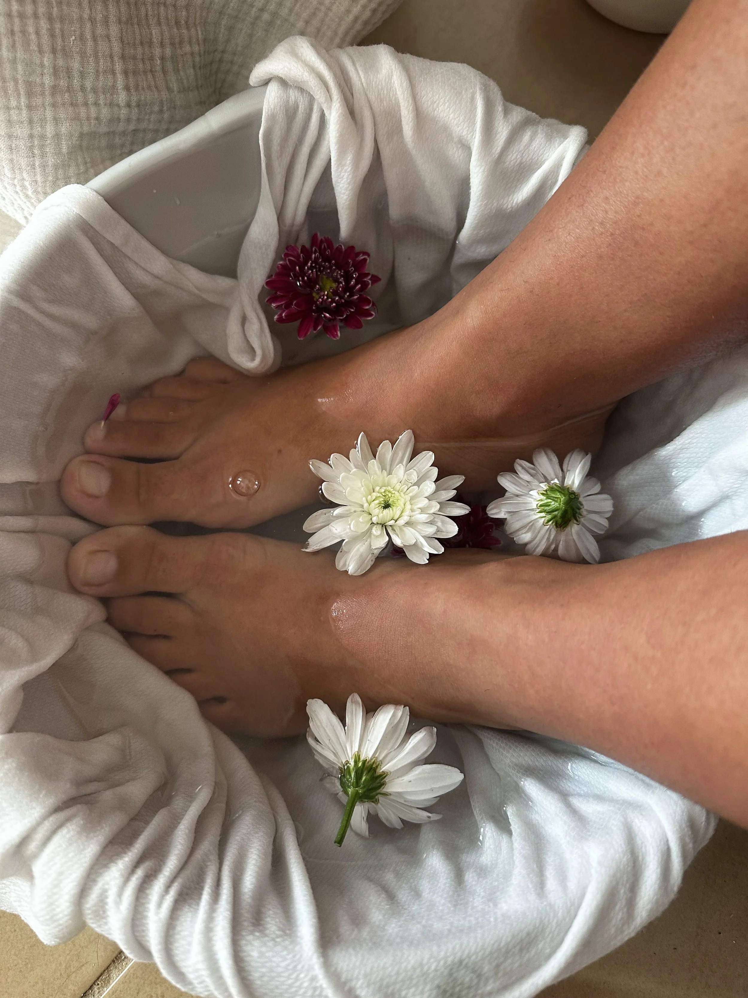 Dos pies sumergidos en agua con flores blancas y una rosa roja rodeados de toallas blancas