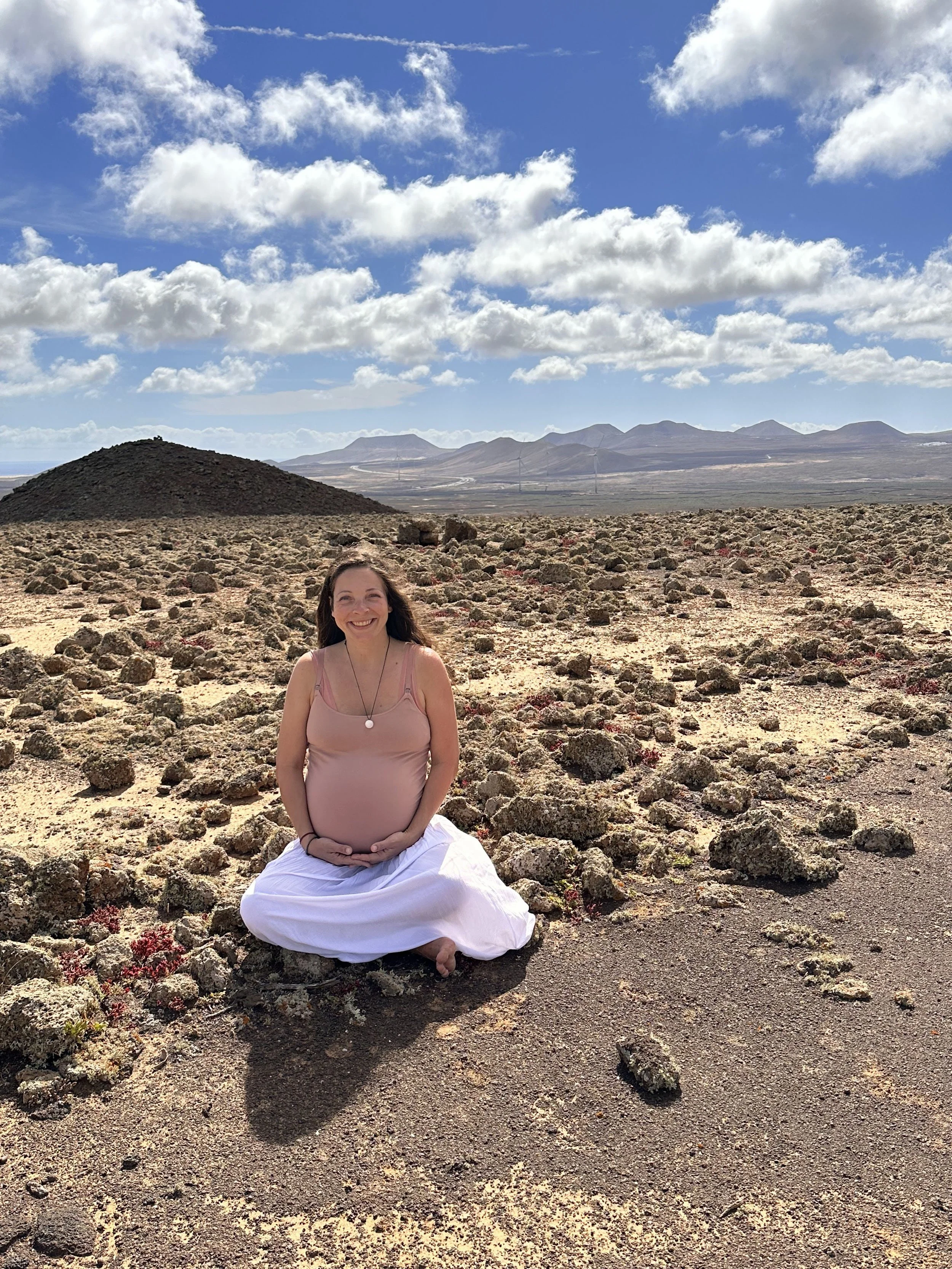 Mujer embarazada sonriendo, sentada en el suelo en un paisaje desértico con rocas, montañas y cielo con nubes.
