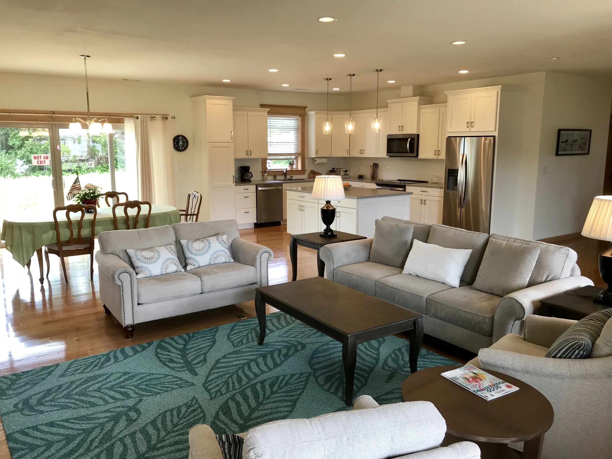 A spacious open-concept living room and kitchen area with beige sofas, a wooden coffee table, a teal leafy-patterned rug, a side table with a magazine, a kitchen with white cabinets, stainless steel appliances, and a dining table with a green tablecloth and wooden chairs. Large sliding glass door with a curtain leads outside.