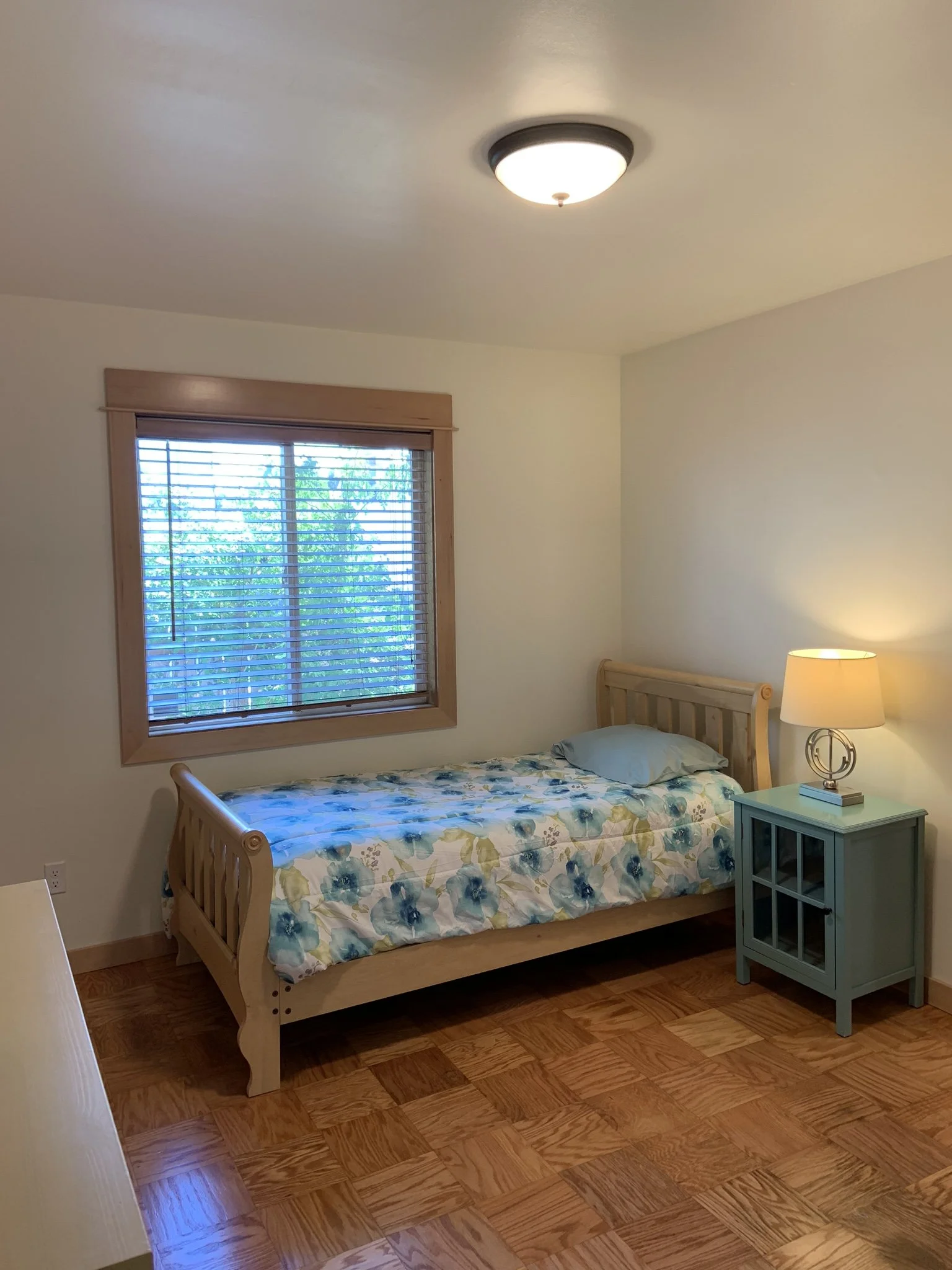 A small bedroom with a wooden bed, a window with blinds, a nightstand with a lamp, and hardwood flooring.