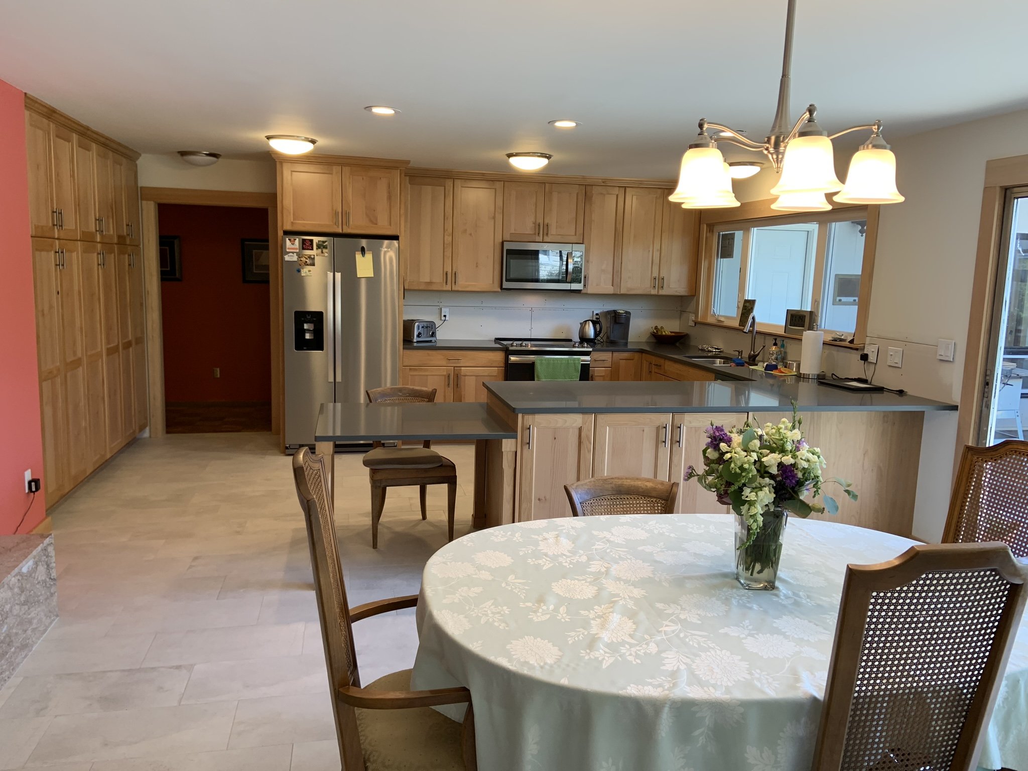 Kitchen with wooden cabinets, a stainless steel refrigerator, microwave, and countertop appliances, a window overlooking the outside, a dining table with a floral tablecloth and a vase of flowers, and a chandelier overhead.