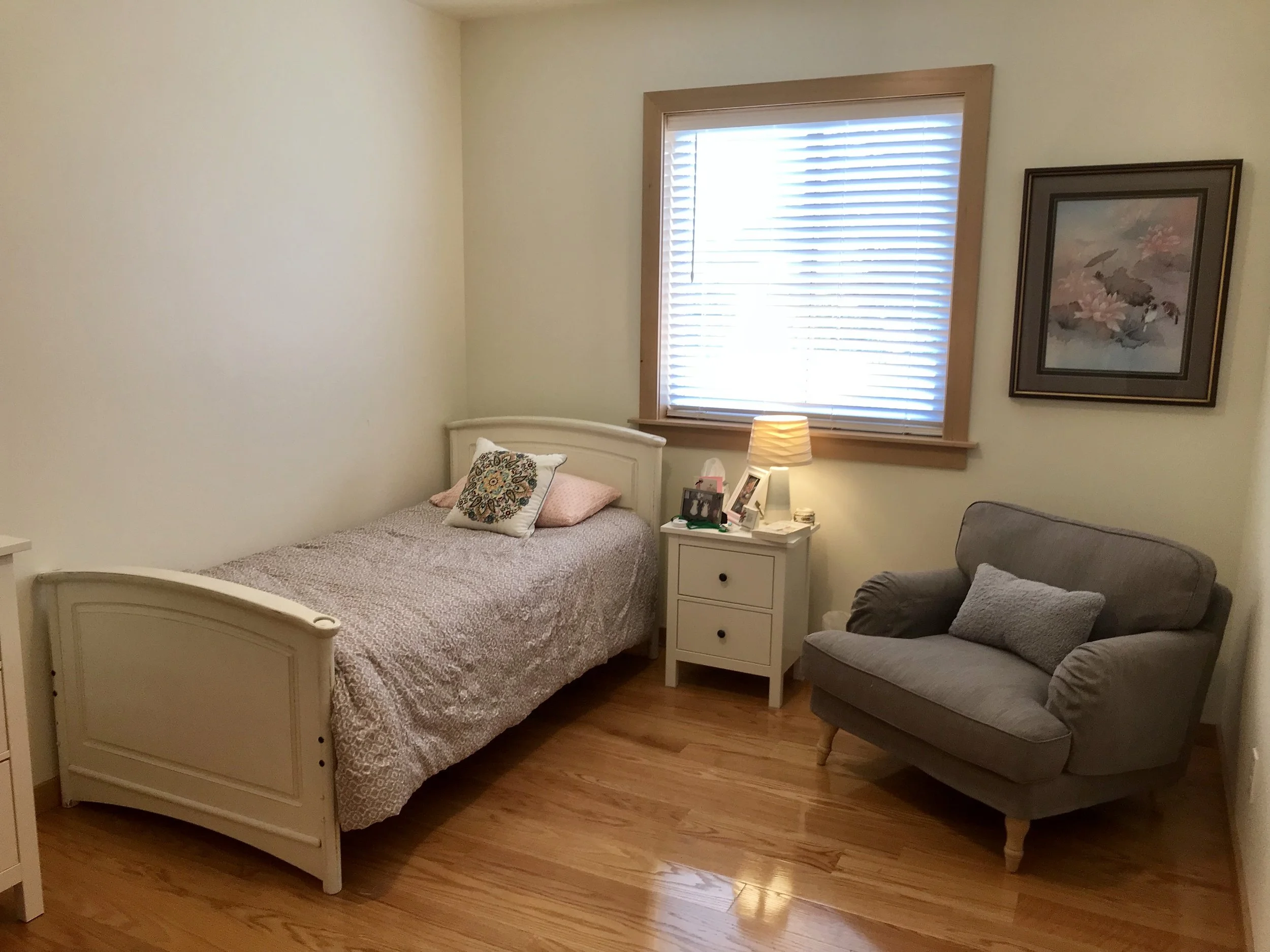 A cozy bedroom with a single bed, colorful pillows, a nightstand with a lamp, framed artwork on the wall, and a gray armchair with a pillow. There's a window with blinds.
