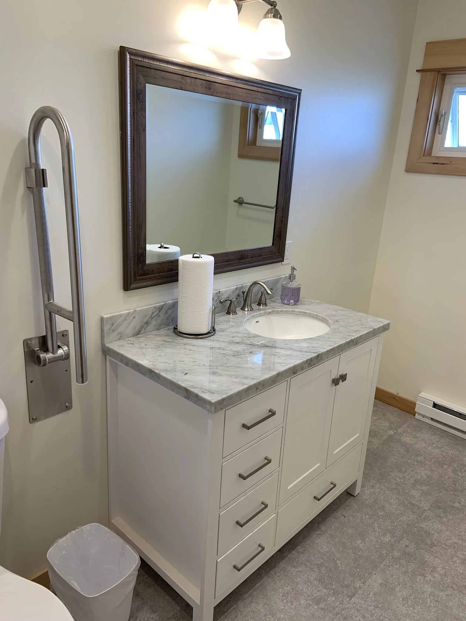 Bathroom with a white vanity, marble countertop, mirror, two small windows with wooden trim, small trash can, and a wall-mounted handrail near the toilet.