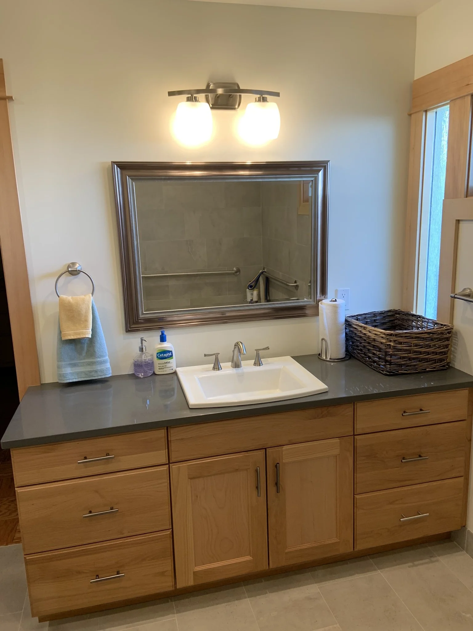 Bathroom vanity with a wooden cabinet, a gray countertop, a white sink, a large mirror, and above it a two-light fixture. Items include soap, hand sanitizer, a face towel, a basket, and paper towels.