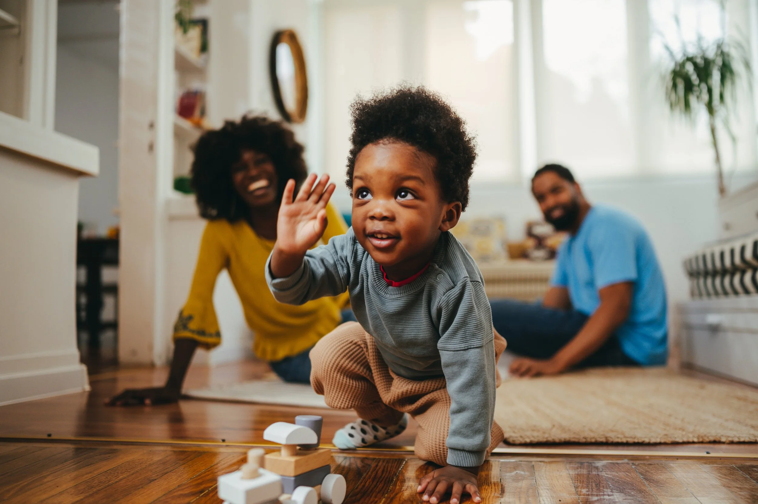 A young boy crawling on the floor, playing with wooden toy blocks, with a woman and man in the background smiling in a bright living room receiving caregiver coaching from a BCBA