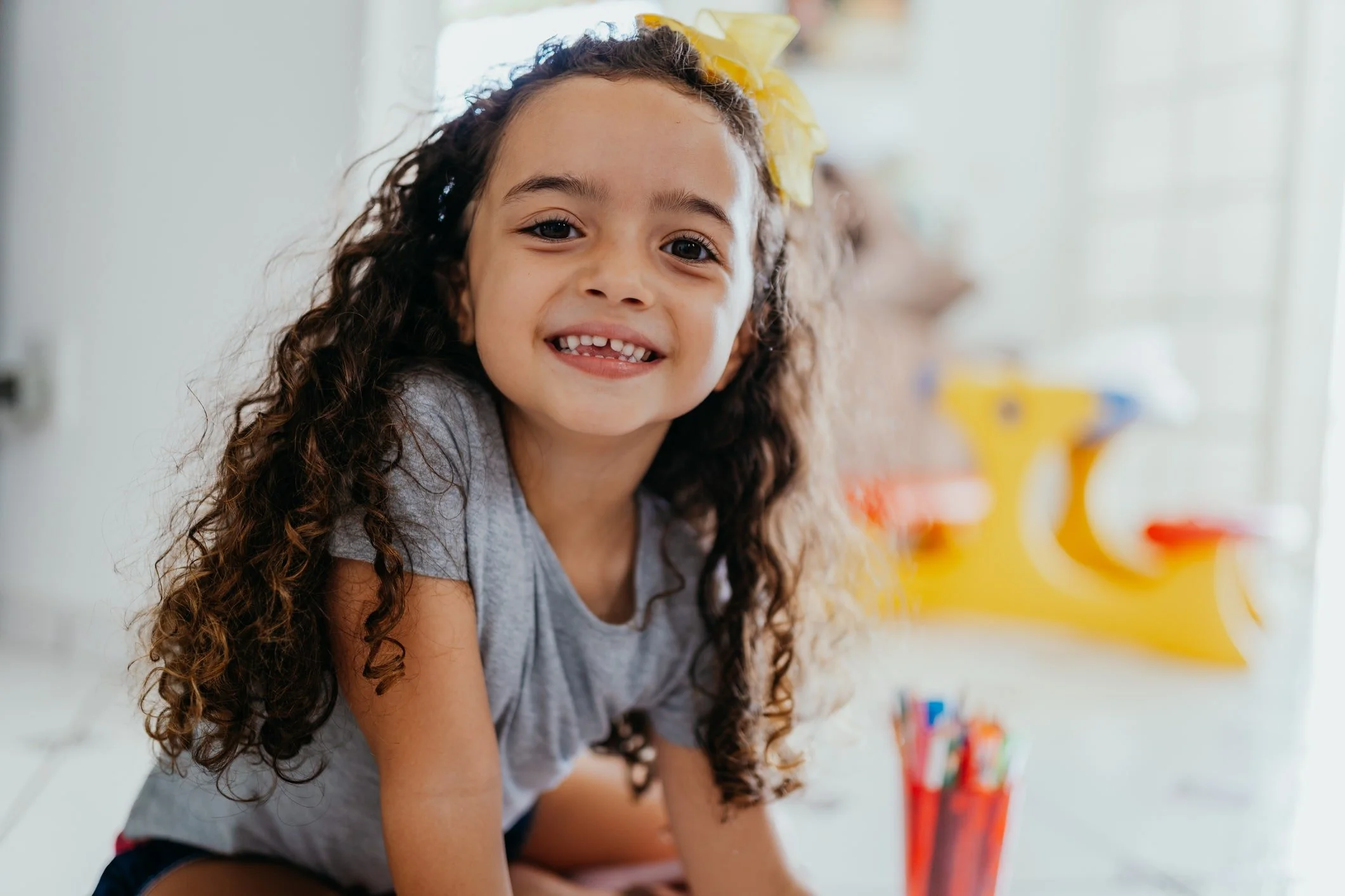 A young neurodiverse girl with long curly brown hair, wearing a yellow bow on her head and a light gray shirt, smiling with missing front teeth, leaning forward in a brightly lit room with colorful toys and books in the background.
