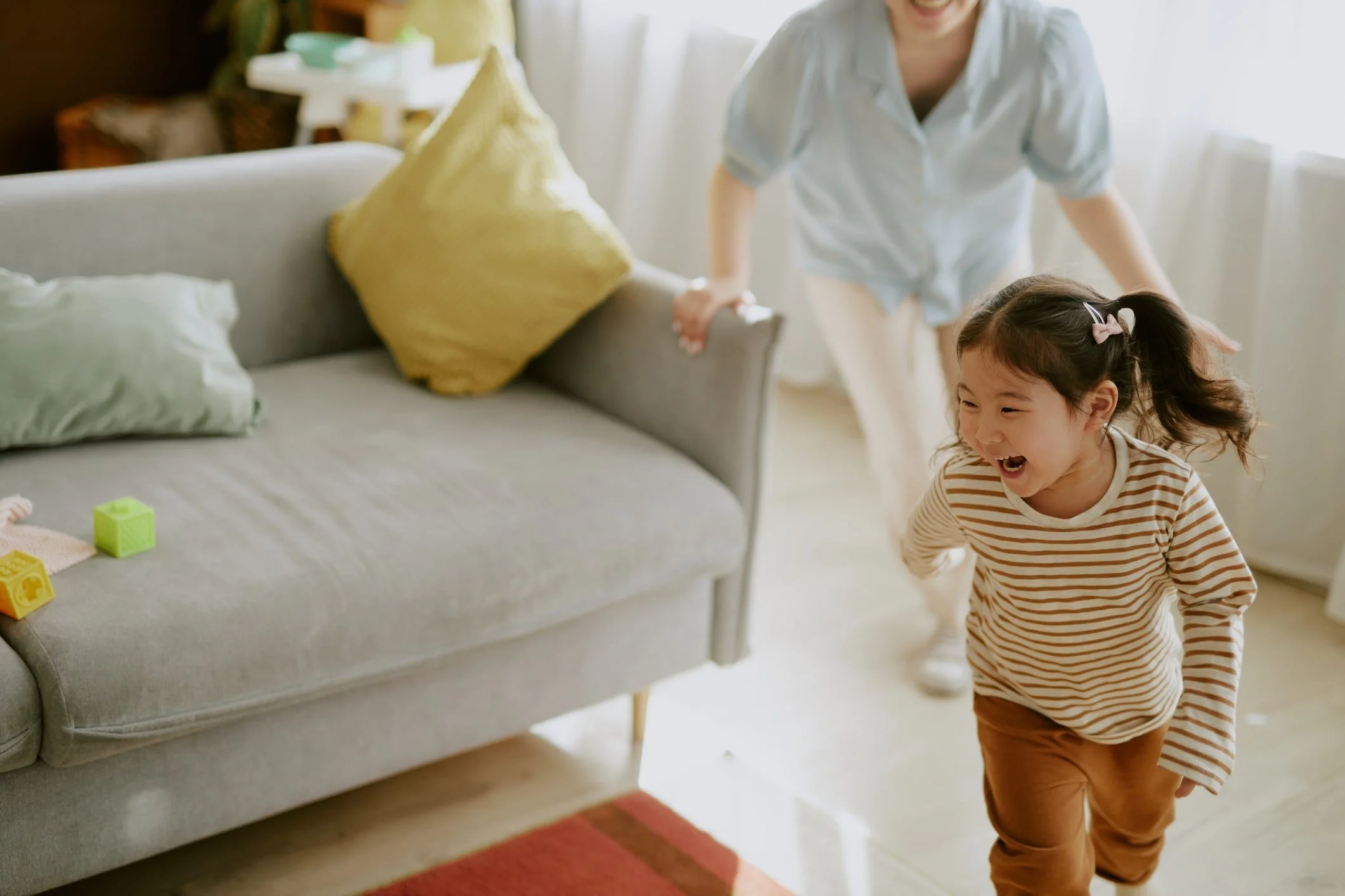 Young girl laughing and running around in a living room with an adult. The room has a gray sofa with yellow pillows and colorful toys on the floor.