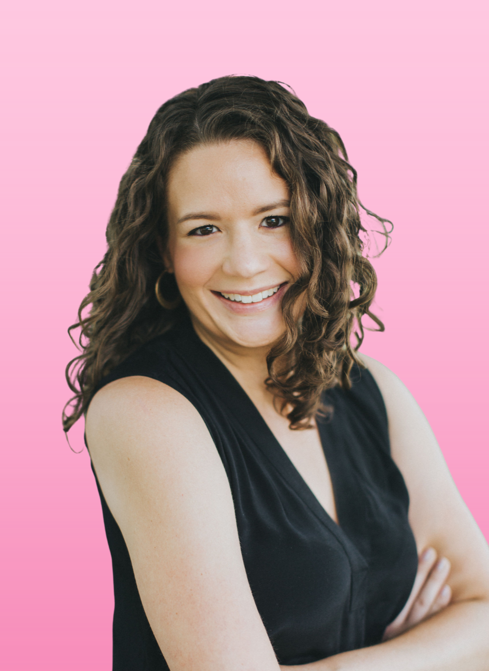 A smiling woman with curly brown hair wearing a black sleeveless top, standing against a pink background.