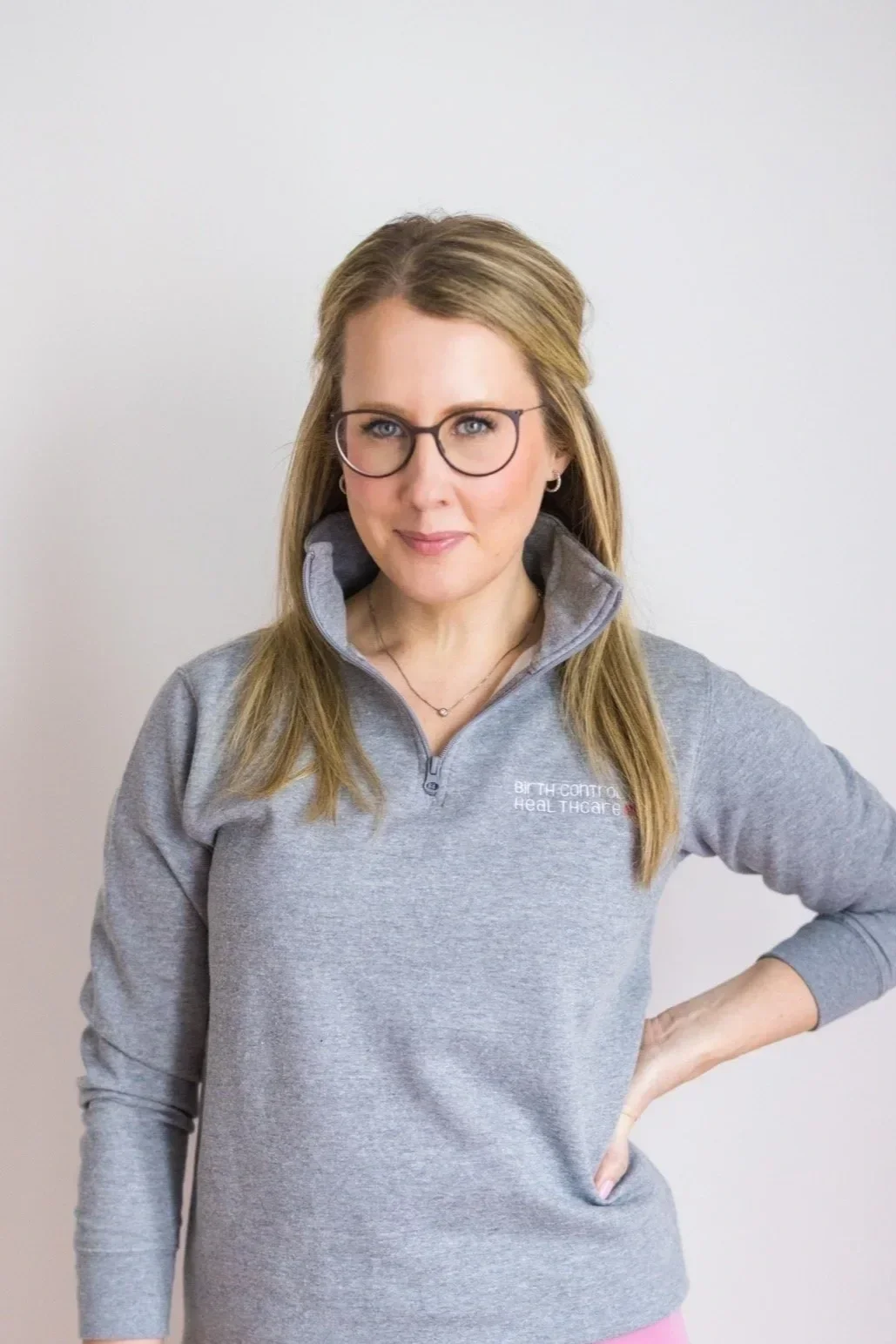 A woman with glasses and light brown hair, wearing a gray zip-up sweatshirt with the words 'Birth Control Healthcare' on it, stands against a plain white background. She has one hand on her hip and is smiling slightly.