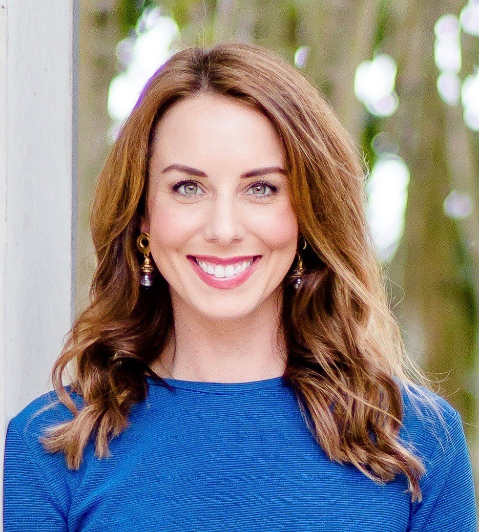 A woman with shoulder-length wavy brown hair, wearing a royal blue top and gold earrings, smiling outdoors with blurred greenery in the background.
