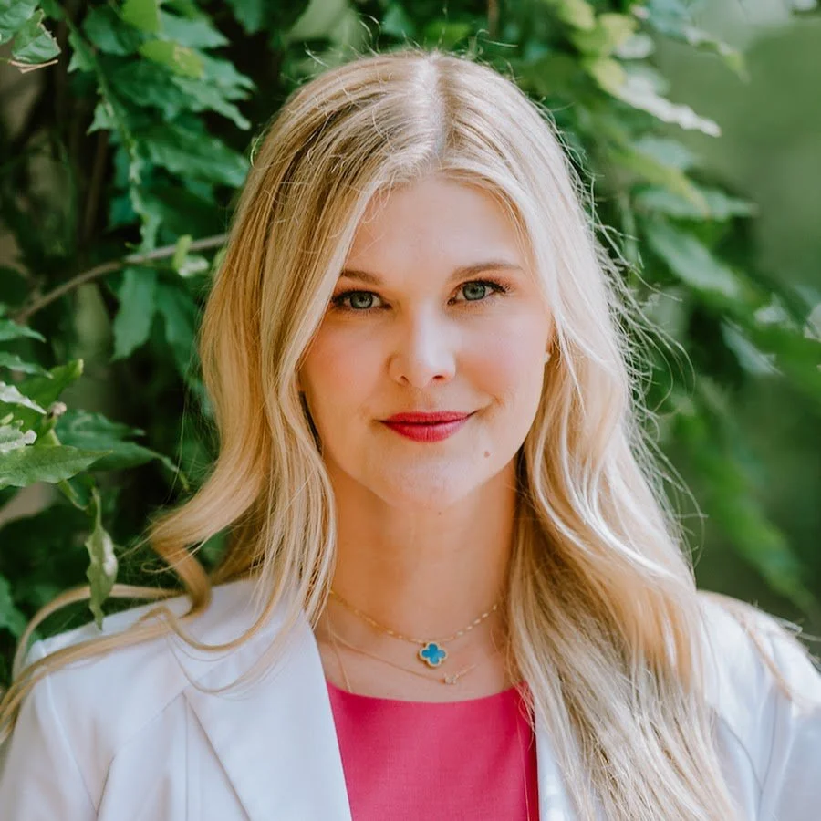 A woman with long blonde hair and blue eyes, wearing a white jacket and pink top, standing outdoors near green leafy plants.