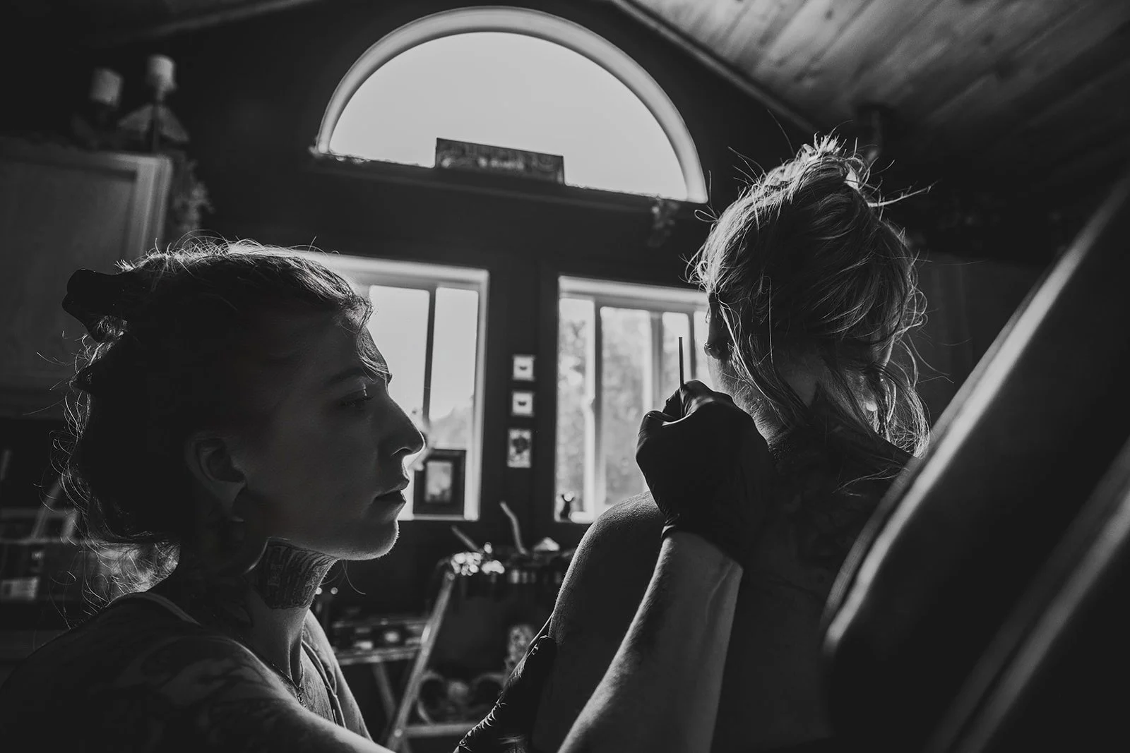 Woman tattooer is tattooing another woman at her private tattoo retreat with large windows and natural light.
