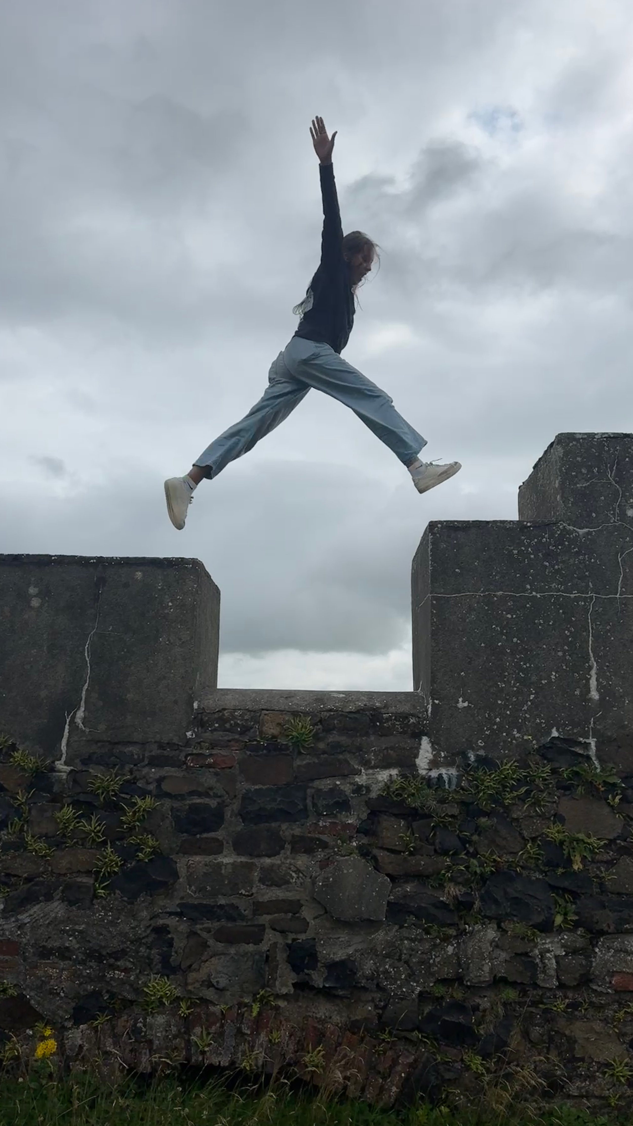 A woman jumping between two large stone structures outdoors on a cloudy day.
