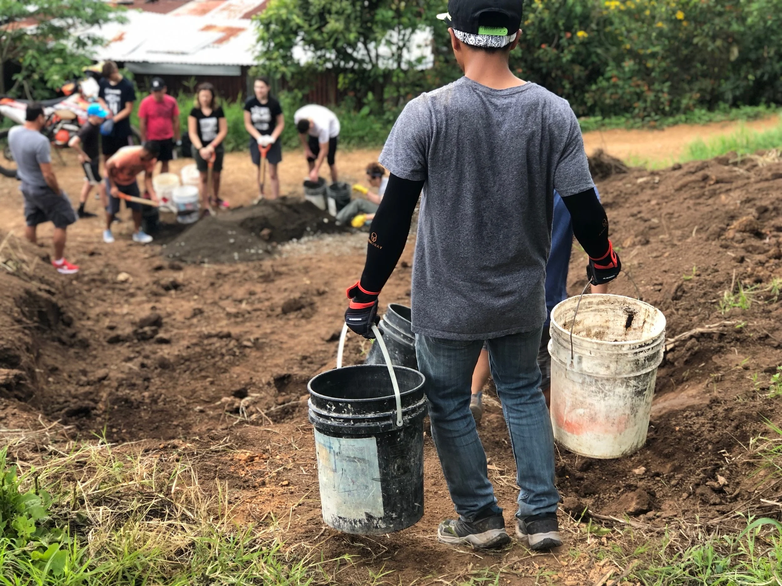 A young man stands outdoors, holding two buckets, watching a group of people working on a community project involving digging and moving soil
