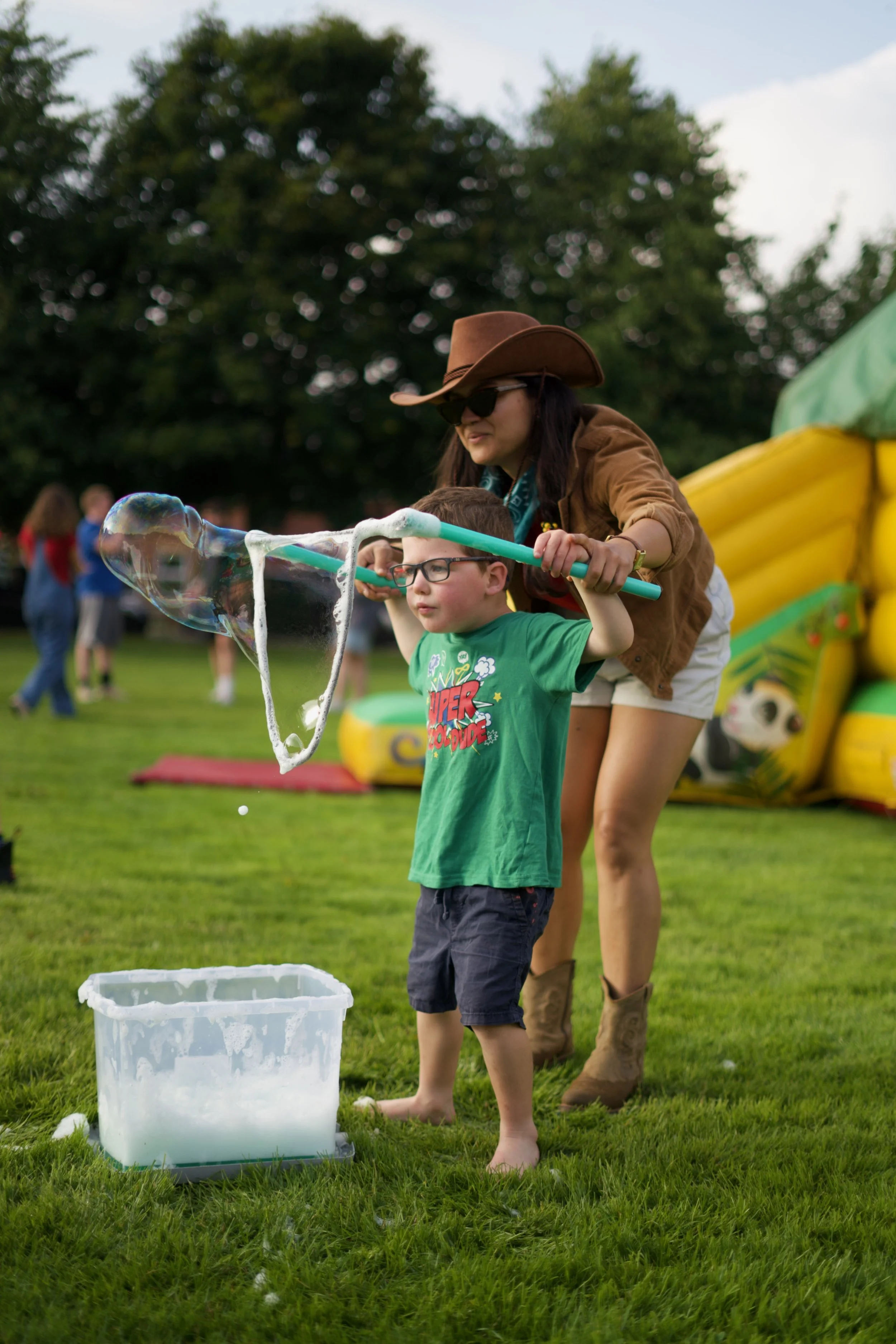 A woman in a cowboy hat and sunglasses helps a young boy in a green shirt blow bubbles using a bubble wand on a grassy field. There are children and people in the background, with a large inflatable bounce house to the right.