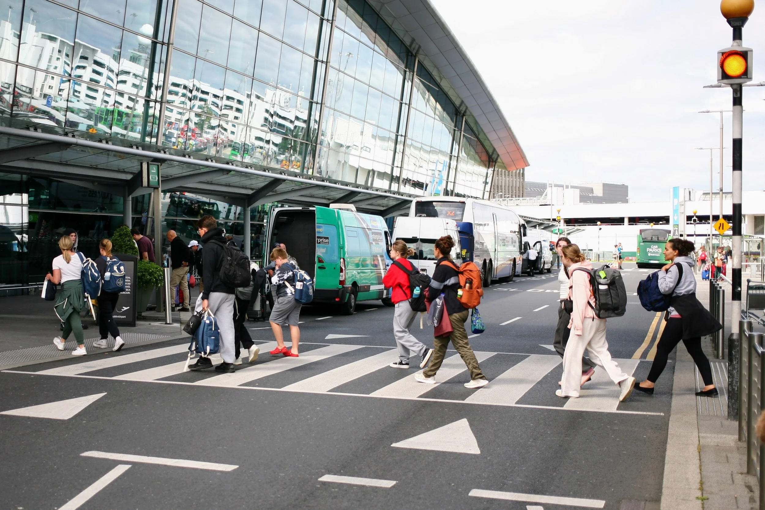 People crossing the street outside an airport terminal with buses and cars nearby.