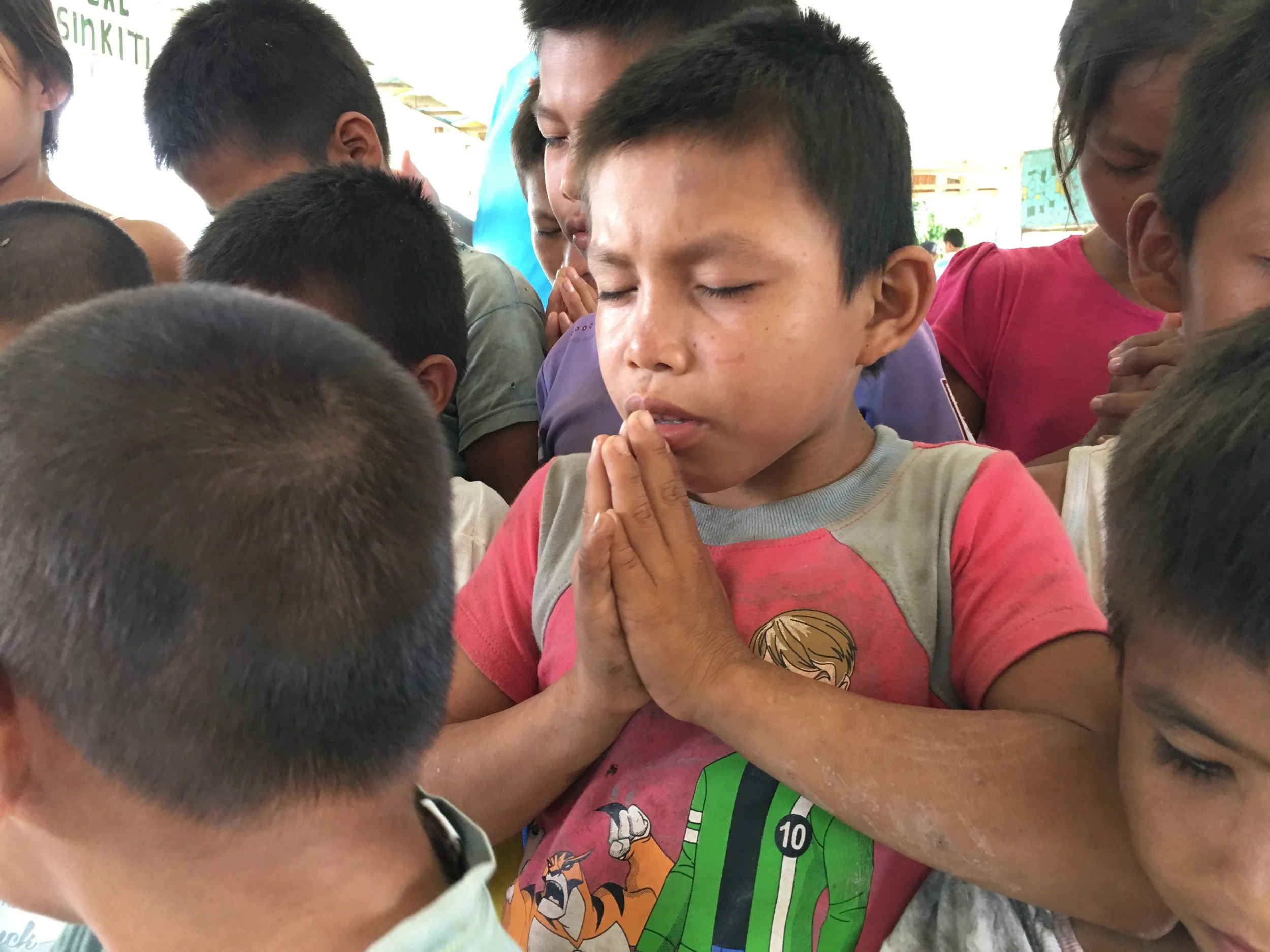 Children in a prayer circle with their eyes closed, hands pressed together, engaged in prayer or meditation.