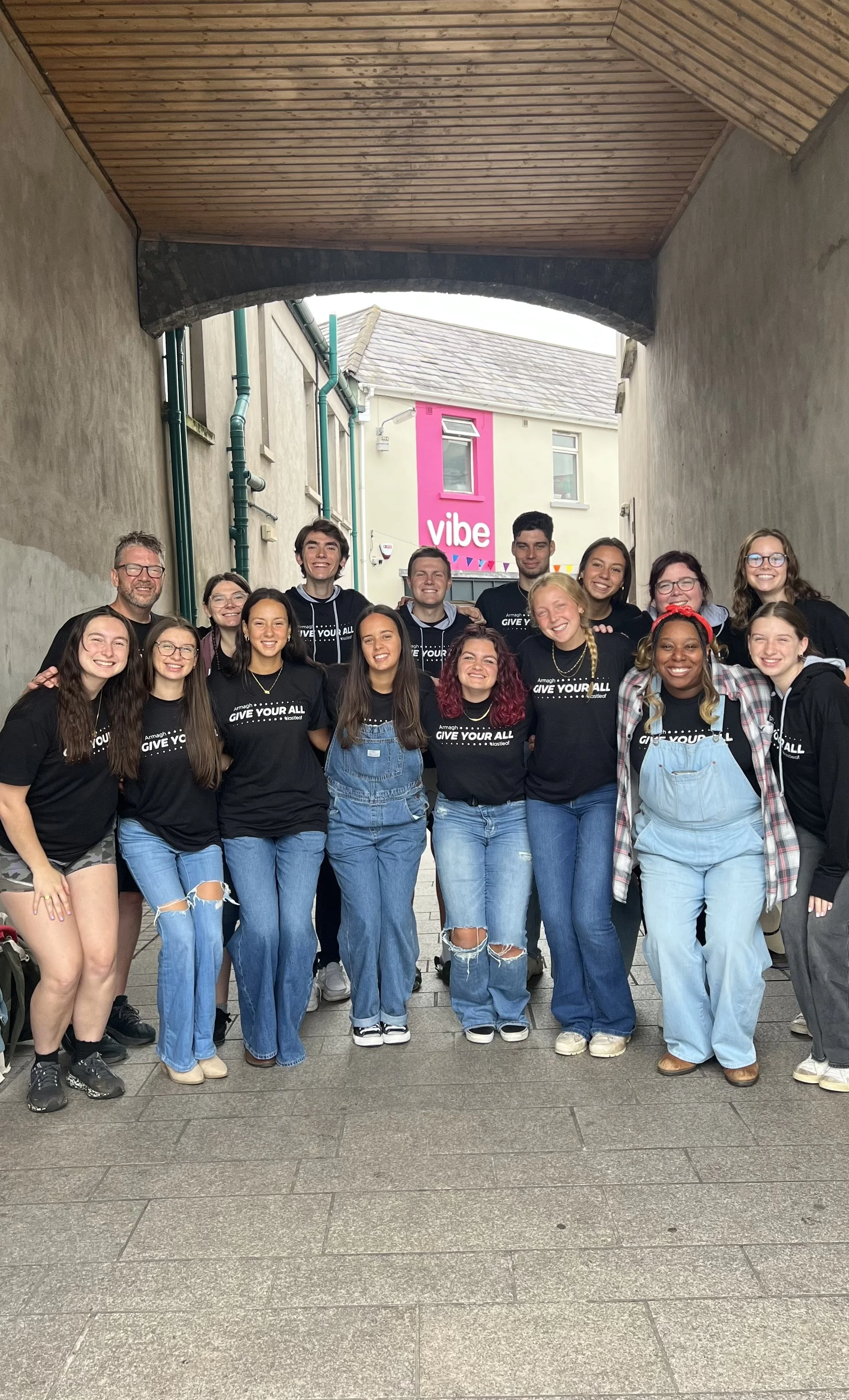 A group of diverse young people and one adult standing together in an alley, smiling for a photo. Most are wearing black T-shirts with the phrase 'GIVE YOUR ALL' printed on them. They are dressed casually, some in jeans, others in shorts or overalls, and they appear to be friends or participants in an event.