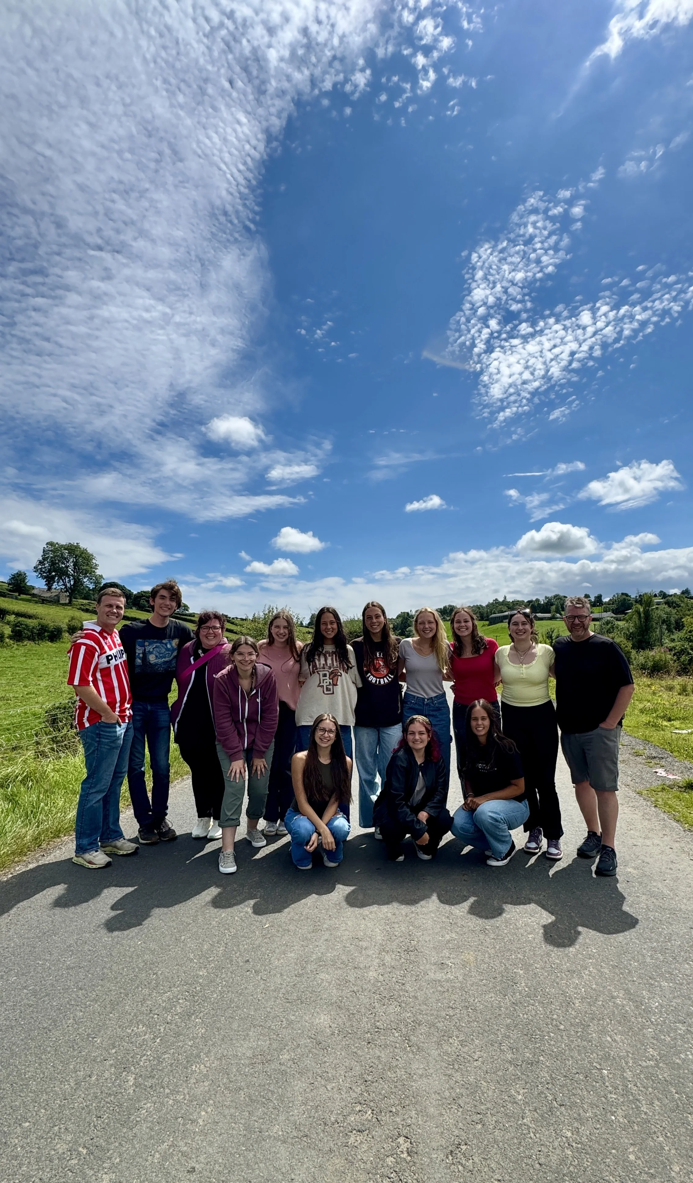 Group of 14 people posing outdoors on a sunny day with a blue sky and scattered clouds, standing on a paved path in a green rural setting.