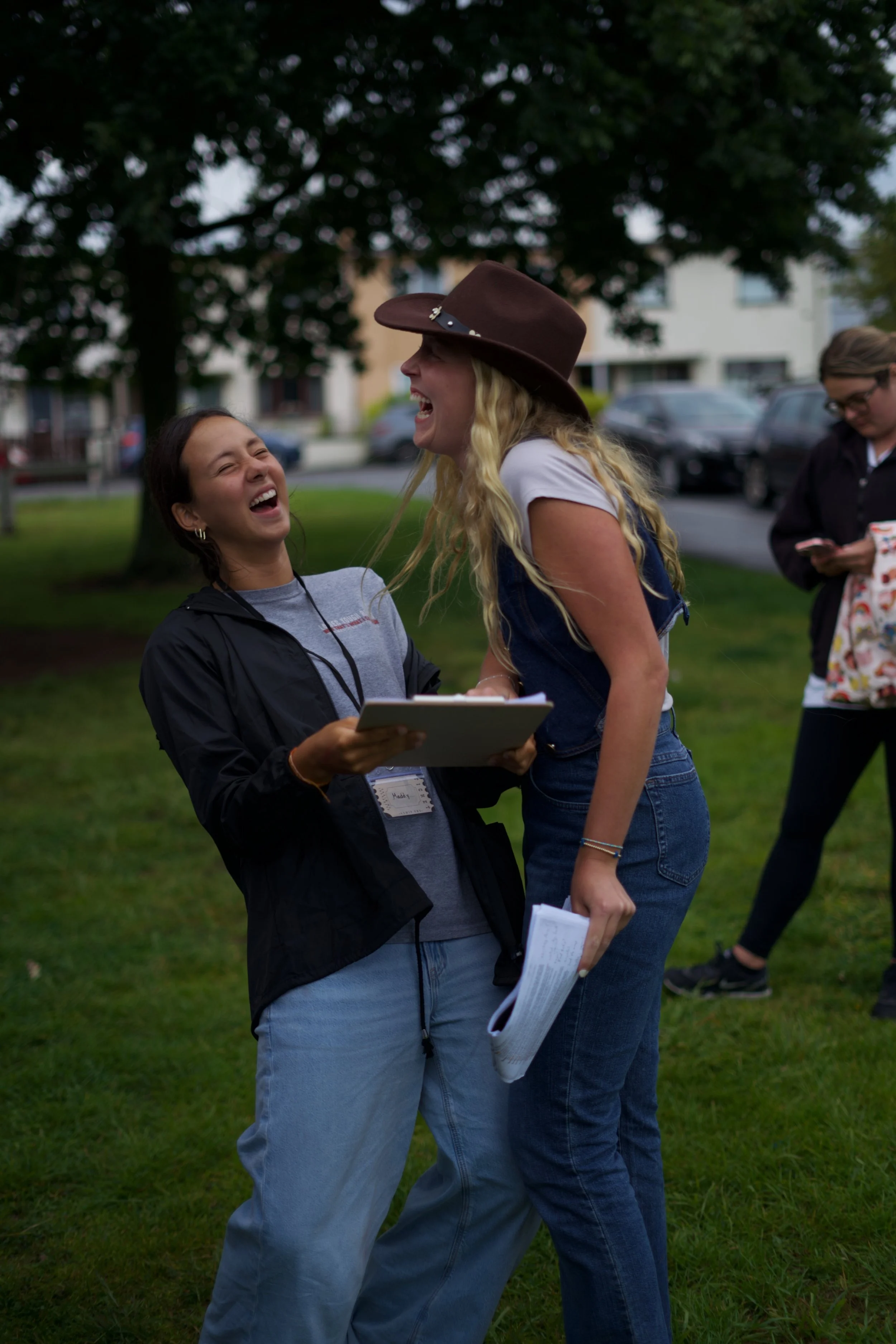 Two women laughing and smiling while talking outdoors on a grassy area, with trees and parked cars in the background.
