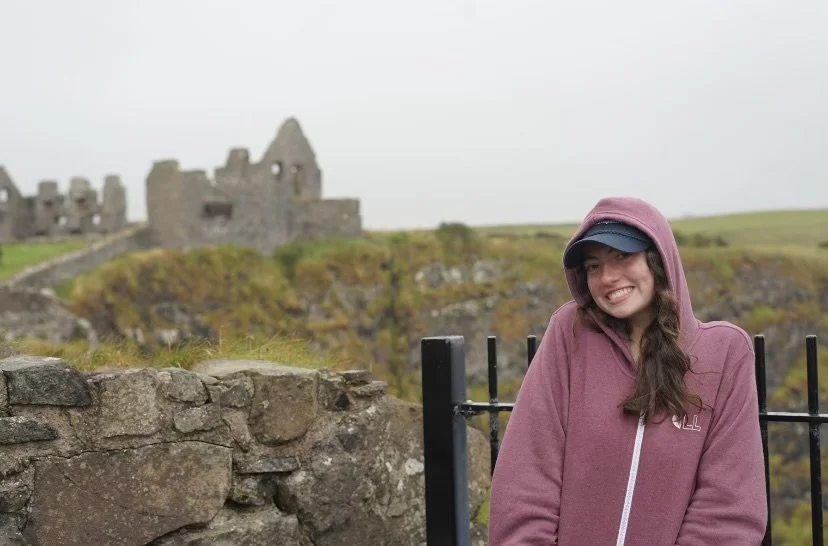Smiling young woman with long brown hair and pink hoodie standing near a stone wall at a historic castle site on overcast day.