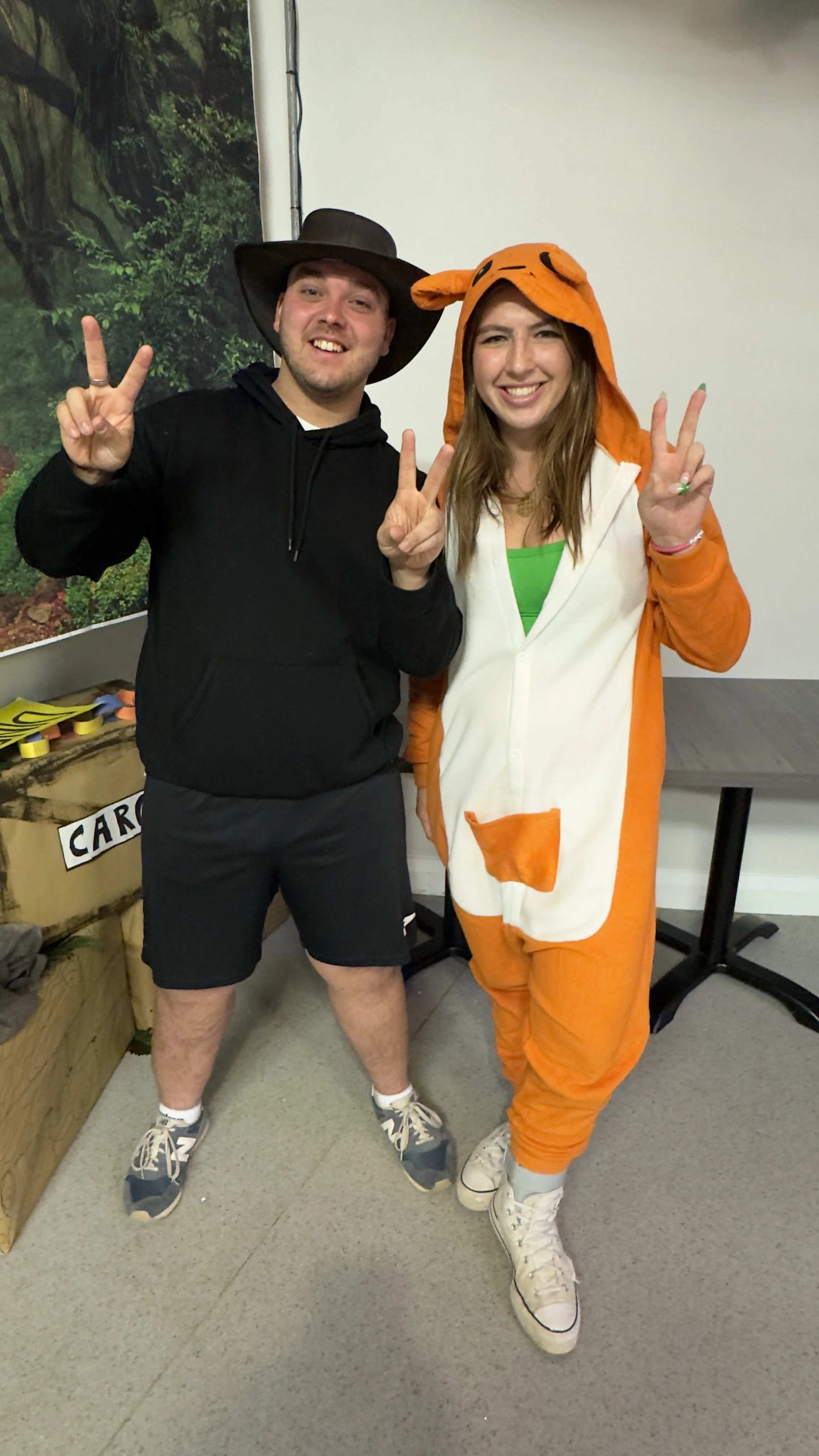 Two people standing indoors, both making peace signs. The person on the left is wearing a large black hat, black hoodie, black shorts, and sneakers. The person on the right is dressed in an orange and white tiger onesie hoodie with tiger ears, tail, and face, paired with white sneakers.