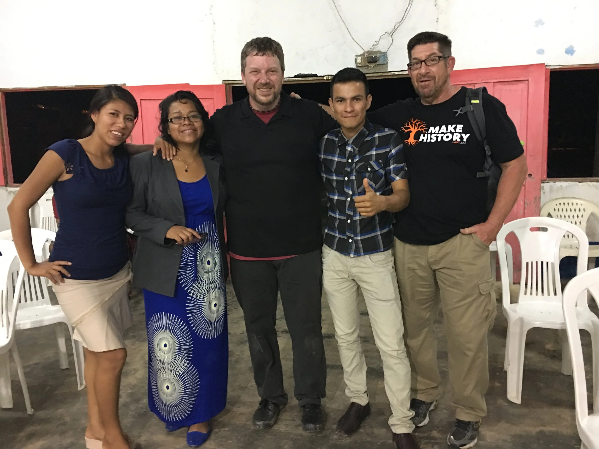Group of five people smiling and posing together indoors, with three chairs visible in the background.