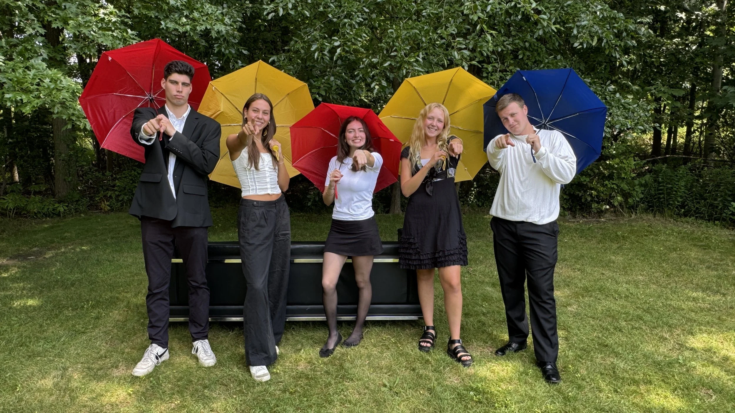 Group of five young people standing outdoors on a grassy area with trees in the background, holding colorful umbrellas and pointing at the camera, smiling and making playful gestures.