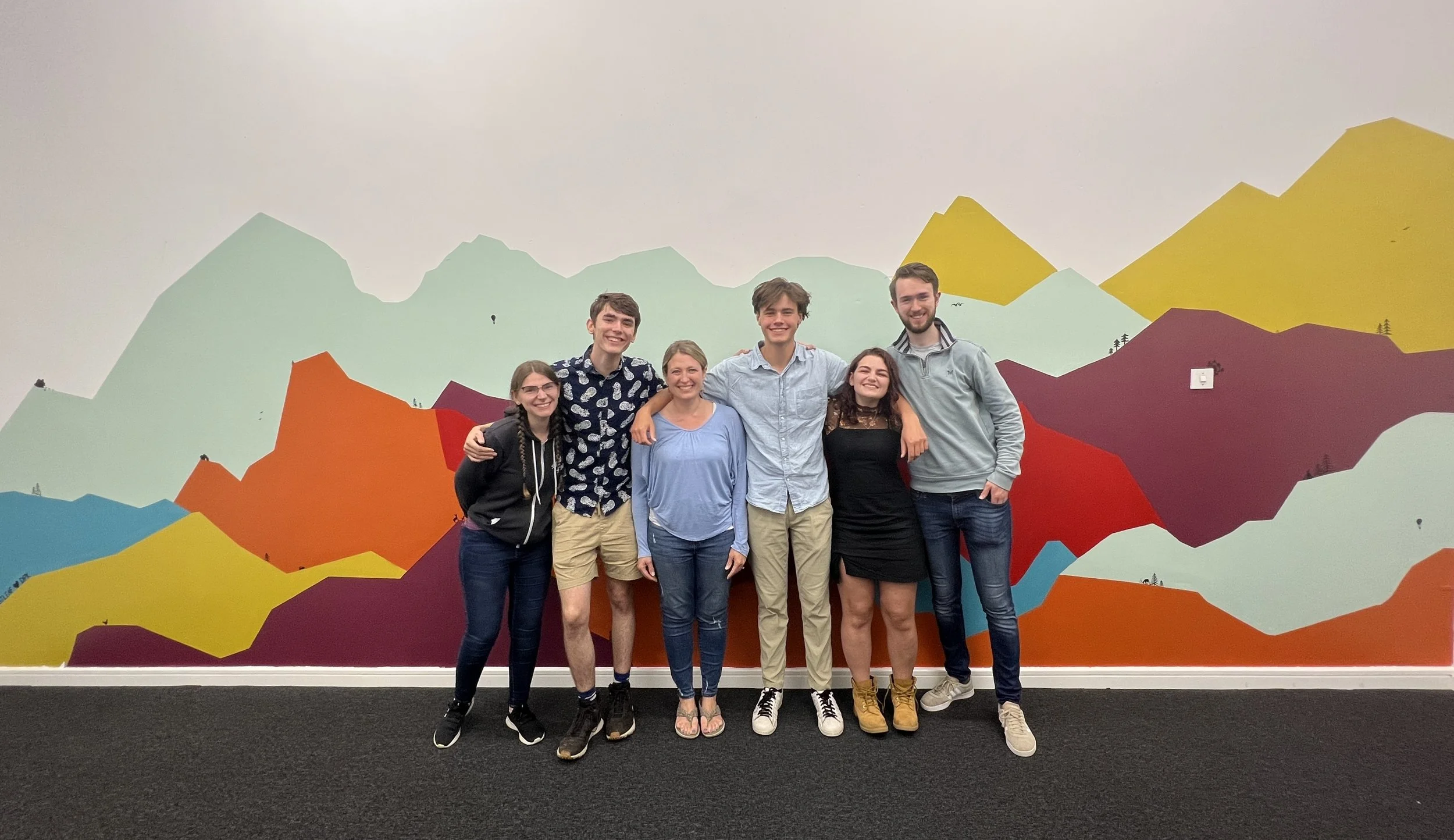 Group of six young adults standing close together indoors, smiling, with a colorful mountain mural mural on the wall behind them.