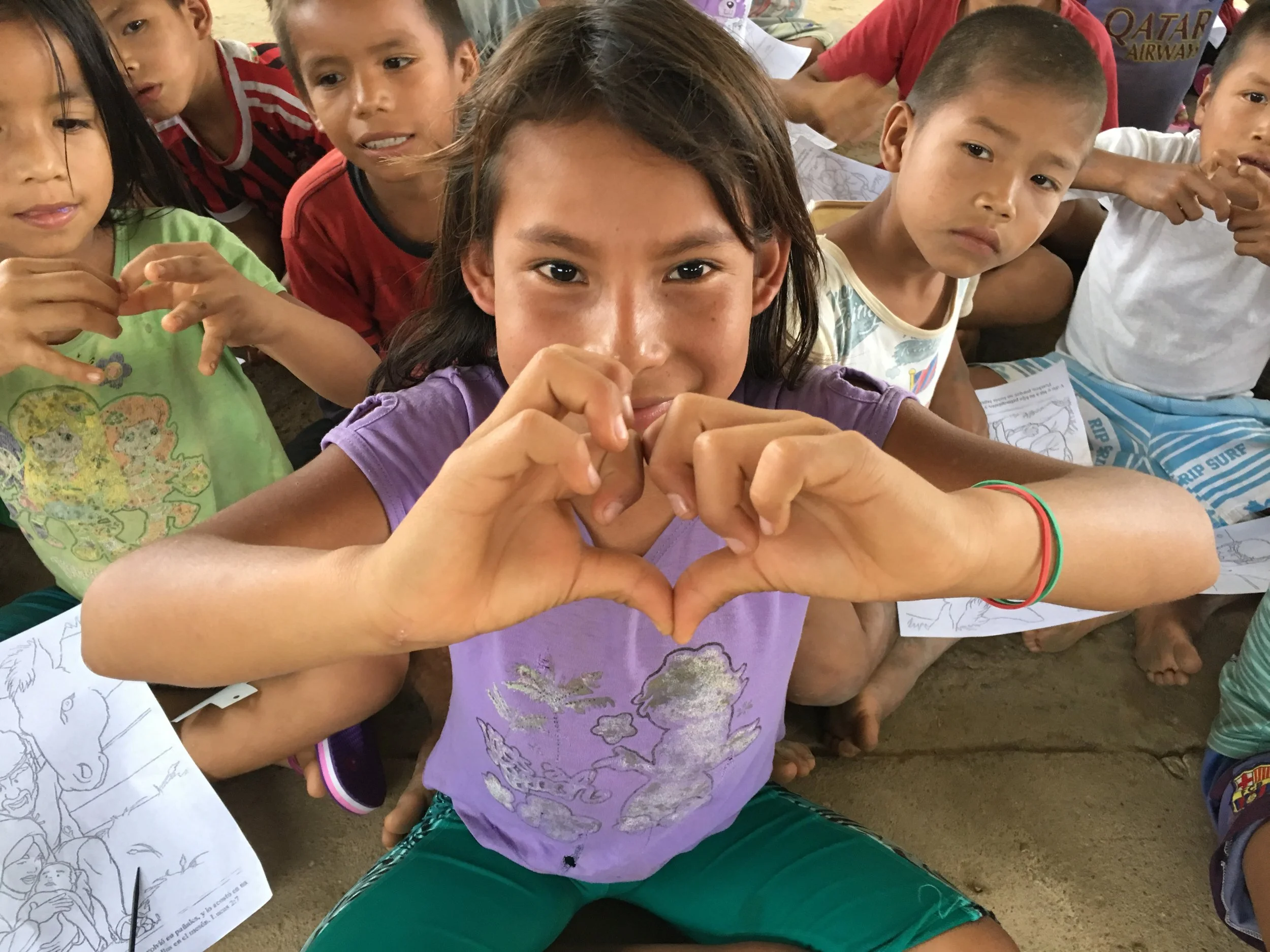 A young girl making a heart shape with her hands, sitting among children with some coloring pages on the ground, appear to be in an outdoor setting.
