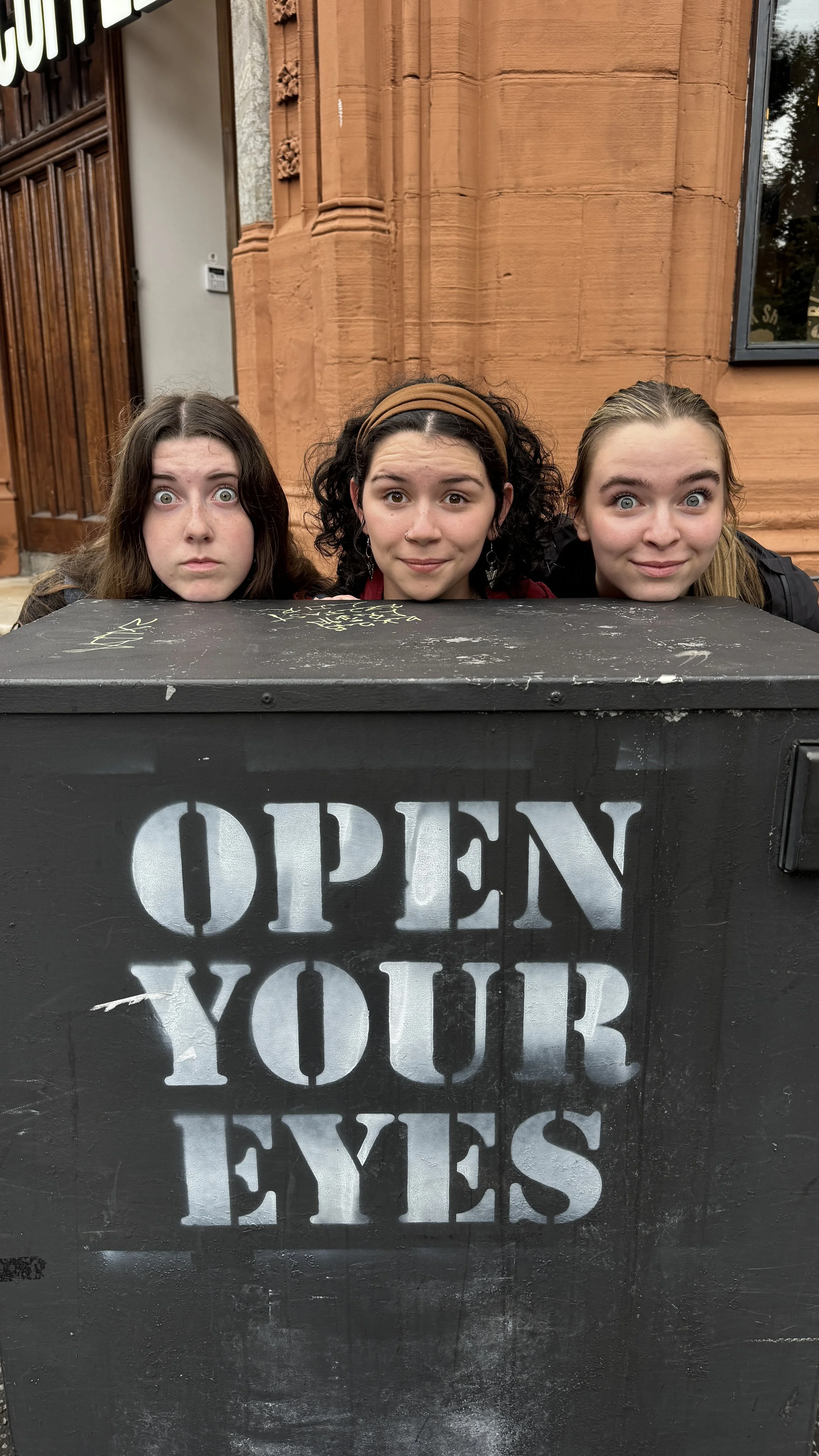 Three young women peering over a black box with the words "OPEN YOUR EYES" painted on it, in front of a brown brick building.