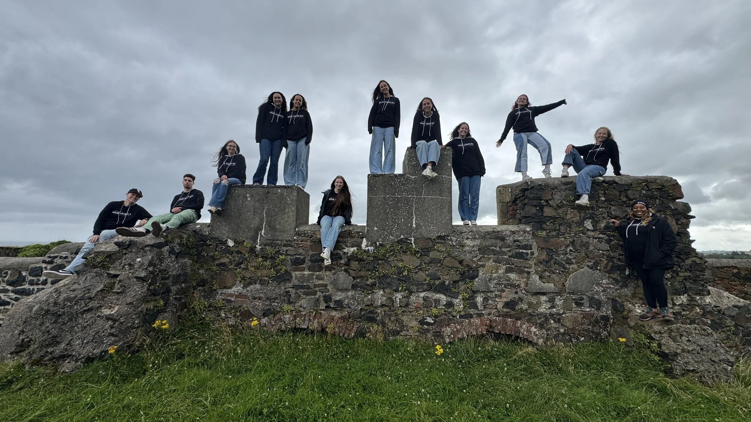 A group of people posing on a historical stone structure outdoors under a cloudy sky, some standing on the stones, others sitting or leaning against the ruins.
