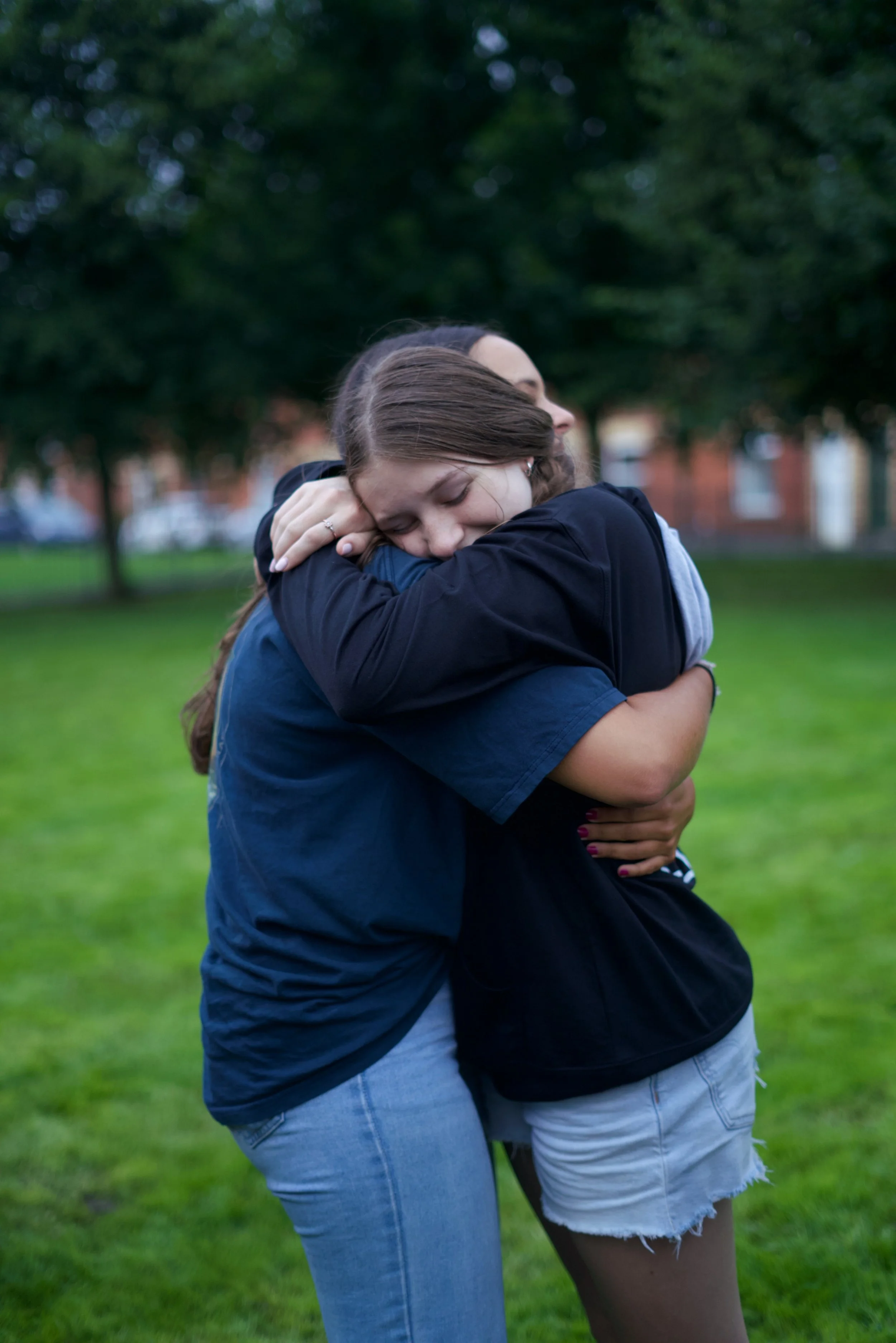 Two women hugging each other tightly in a park with green grass and trees, showing emotional comfort.