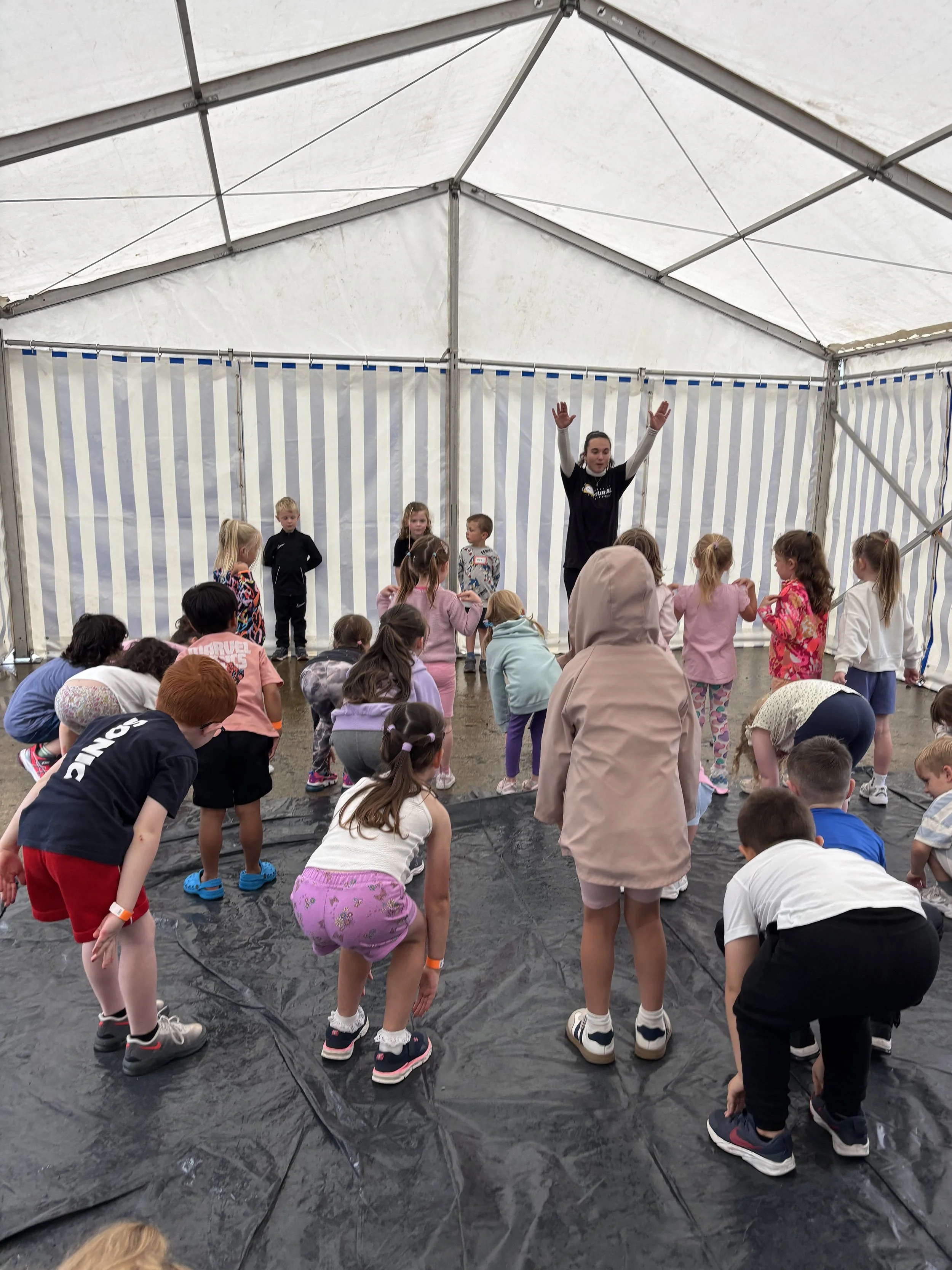 A group of children participating in an activity inside a large tent with white and gray striped walls. An adult instructor stands at the front with arms raised, leading the children, some of whom are bending down, as part of a playful or exercise activity.
