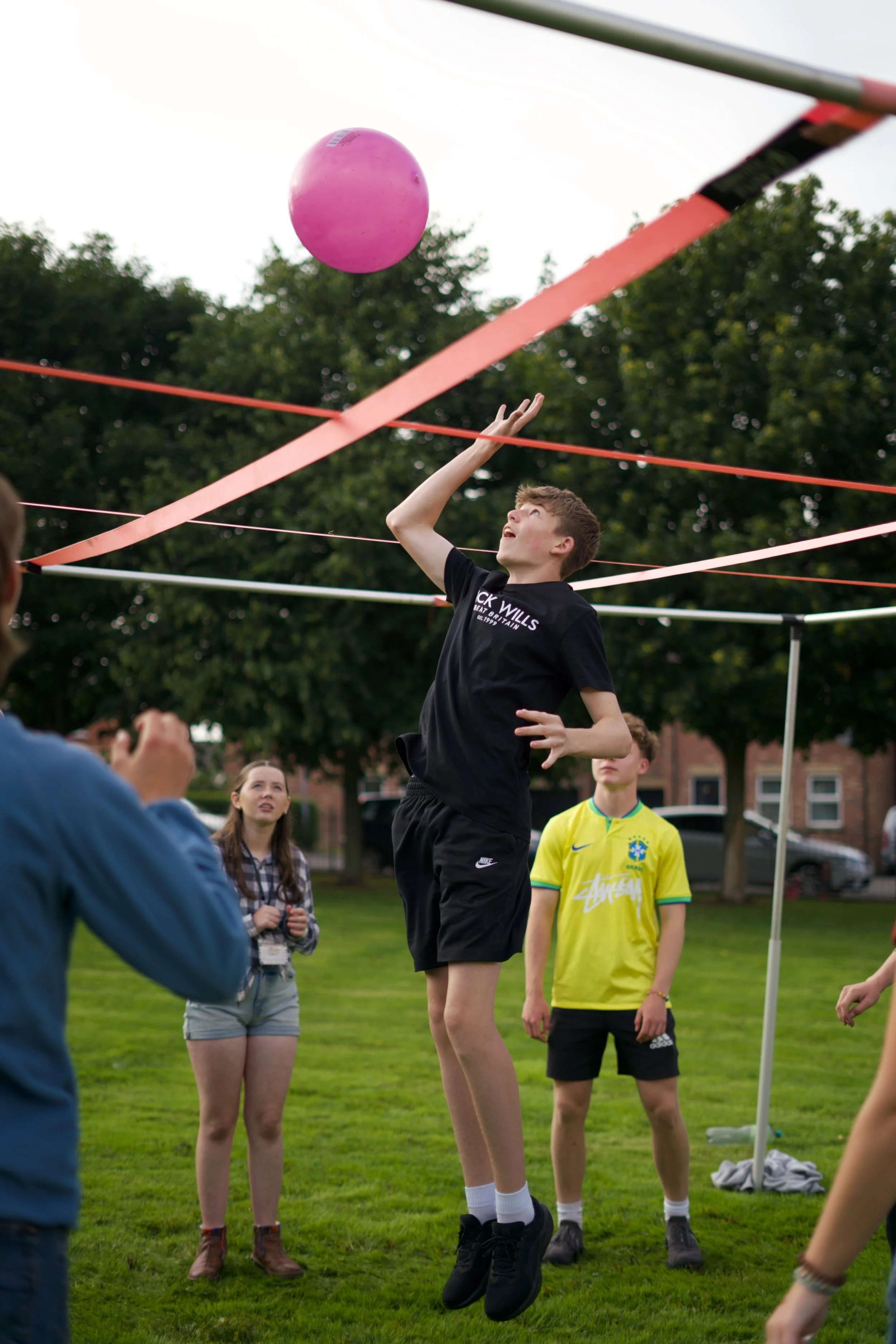 A young boy jumping to hit a pink volleyball on a volleyball net in an outdoor park, with spectators watching.