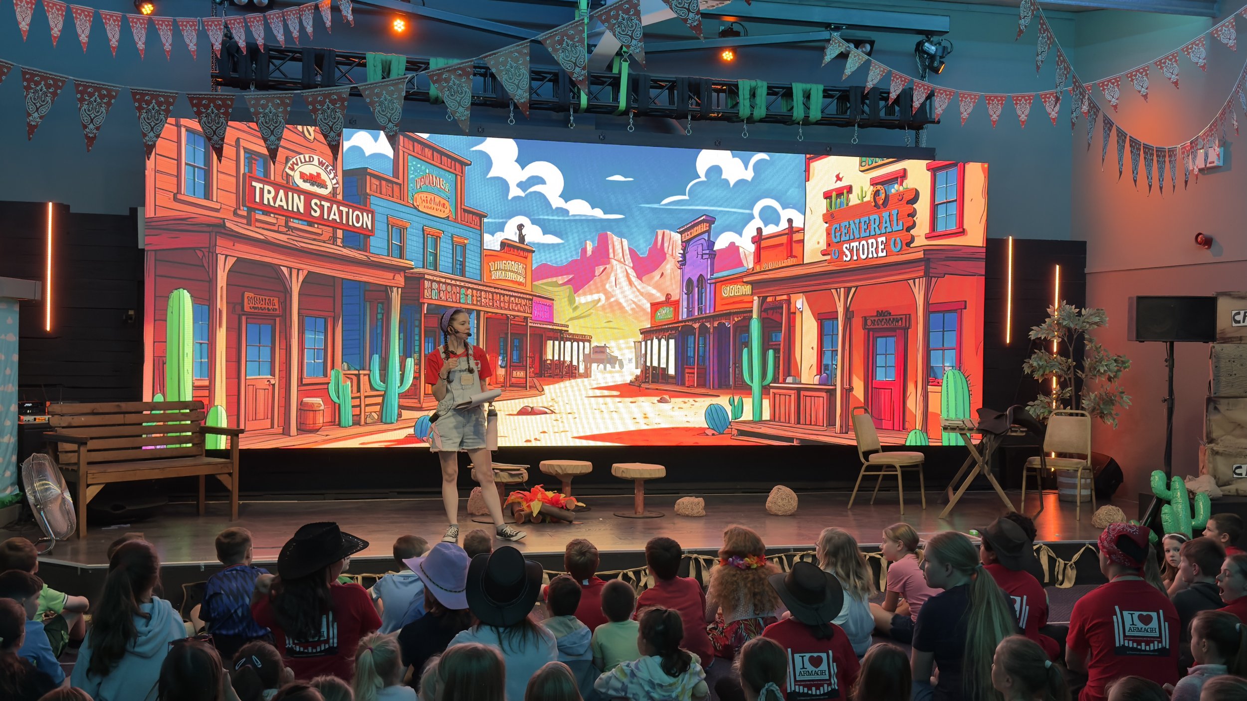 A stage set with animated Western town backdrop, including buildings labeled 'Train Station' and 'General Store,' and desert landscape with mountains, cacti, and rocks, where a performer and children are seated in an audience.