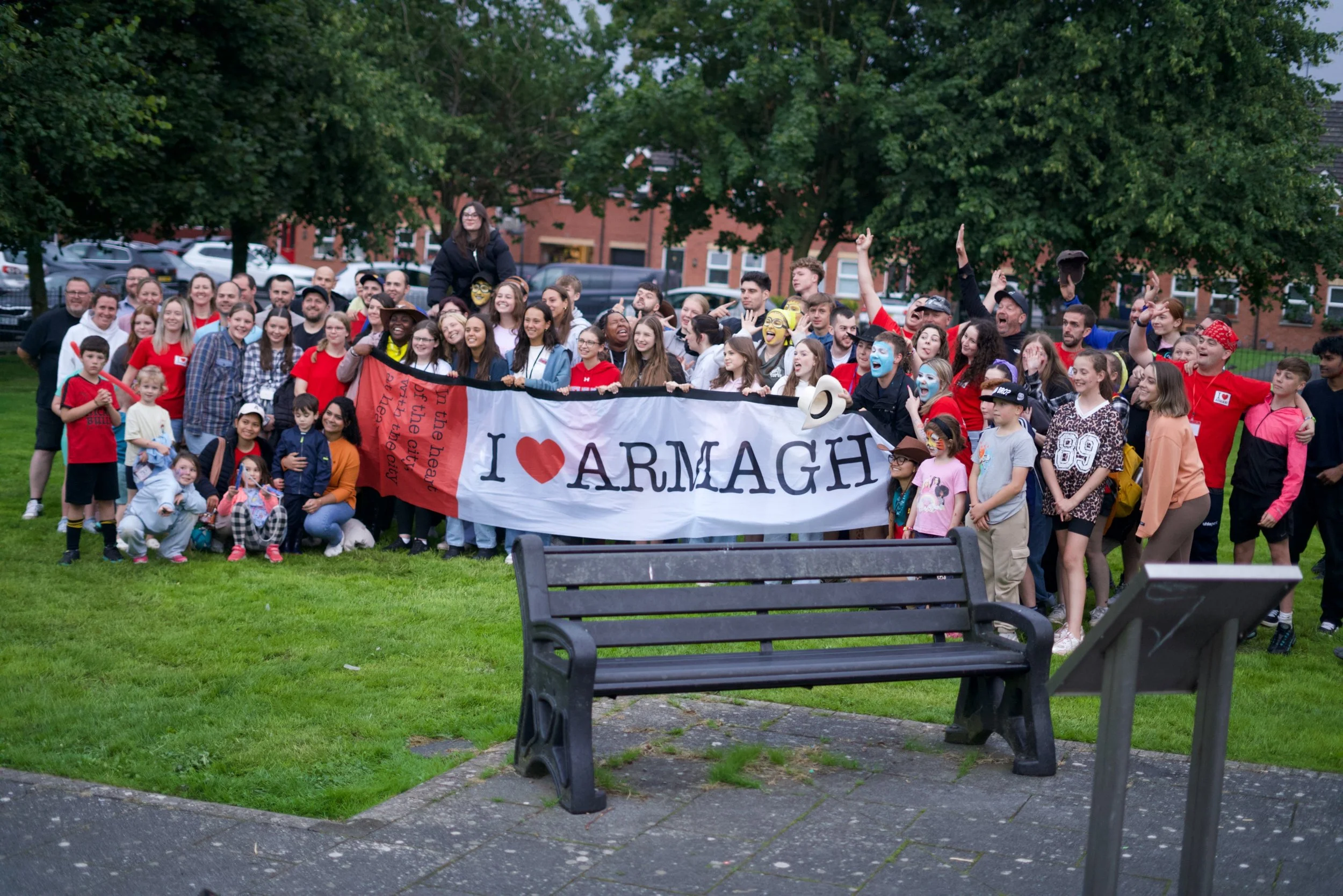 Group of people holding a large banner that reads 'I ❤️ ARMAGH' during outdoor event in a park with green grass, trees, and residential buildings in the background.