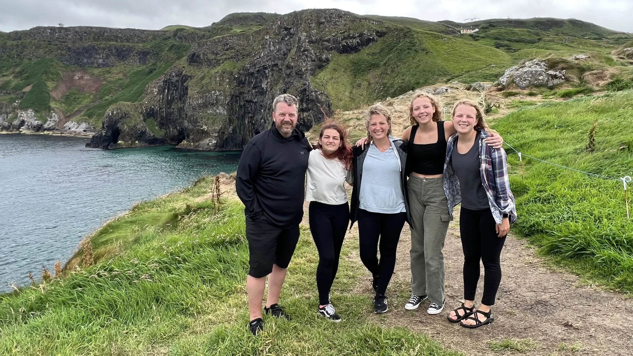 Five people standing on a grassy trail next to a body of water with green hills and rocky cliffs in the background.
