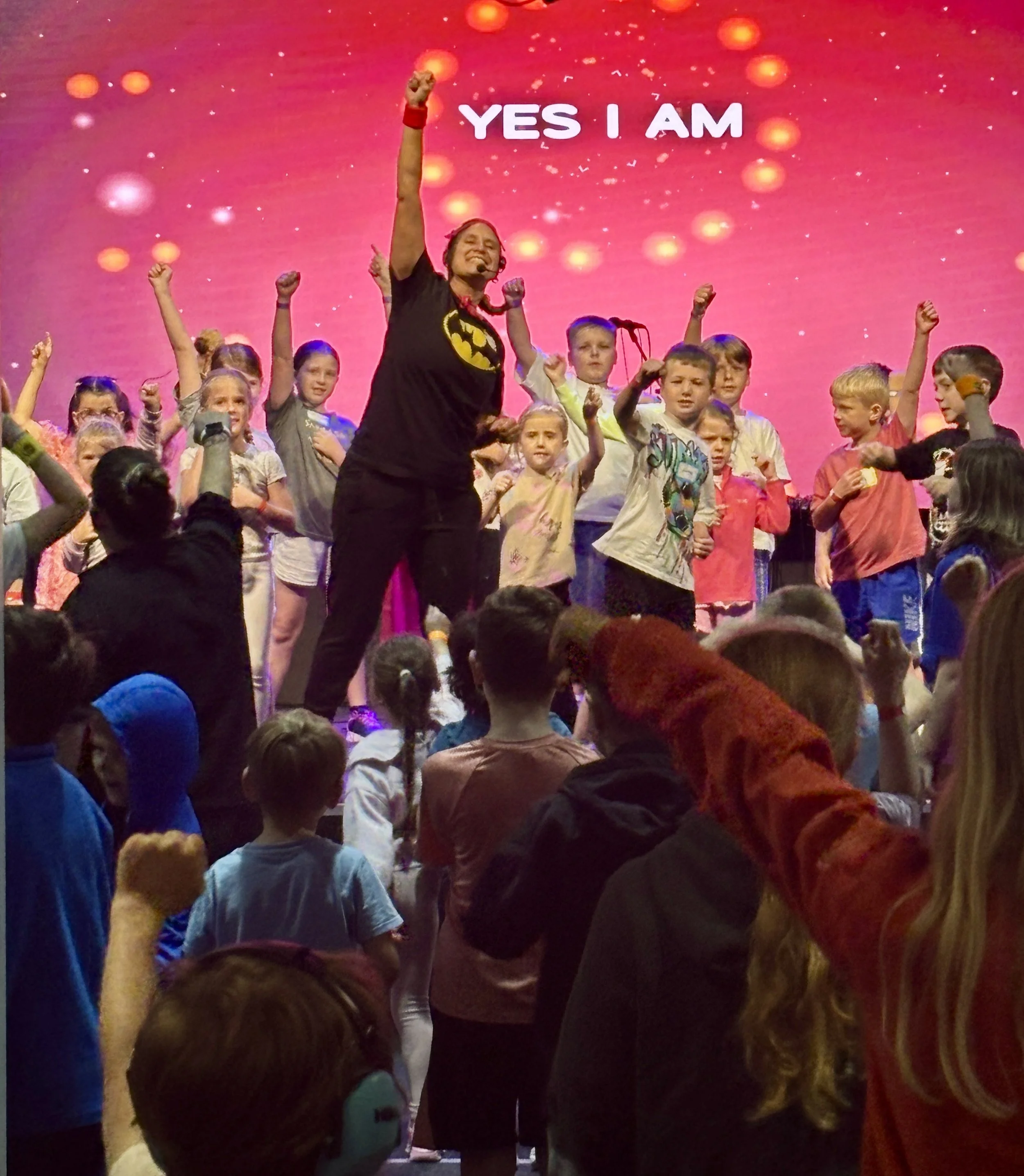 A woman leading a group of children on stage during a performance, with the backdrop displaying the words "YES I AM." The audience is standing and raising their fists in the air.