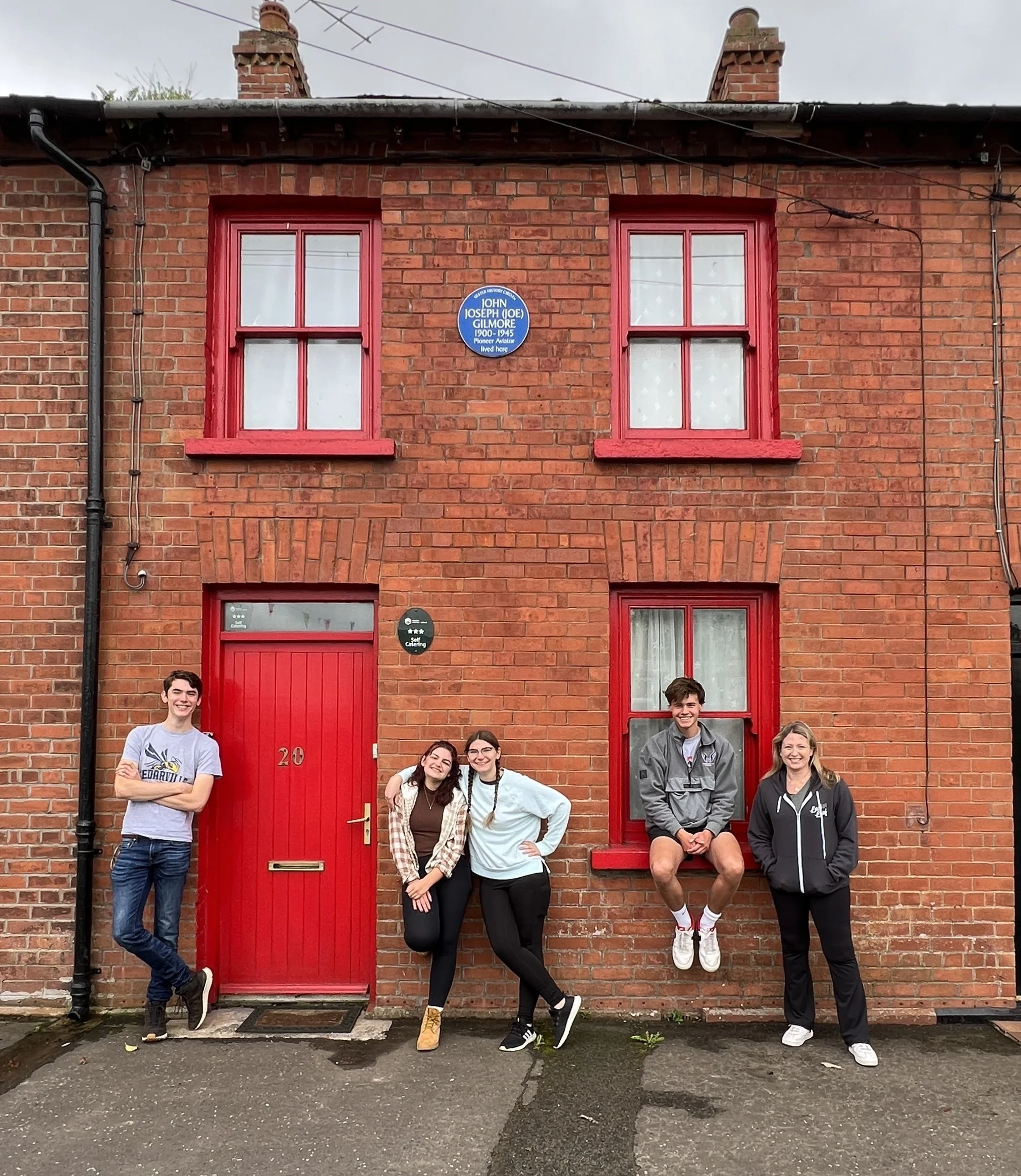 Five people posing outside a brick building with red door and window frames. A man with crossed arms is standing on the left, two women are in the middle, sitting and leaning, a young man is sitting on the window ledge, and a woman is standing on the right. The building has a blue plaque and a red door marked with the number 20.
