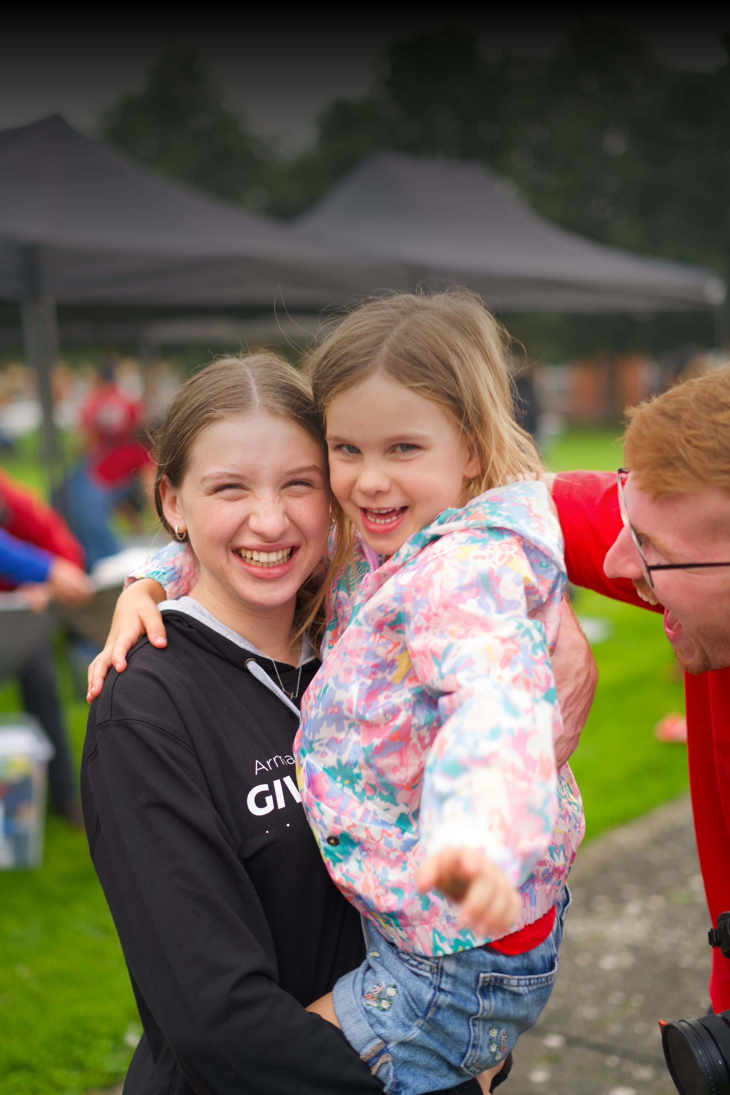 Two young girls smiling and hugging at an outdoor event with tents and people in the background.