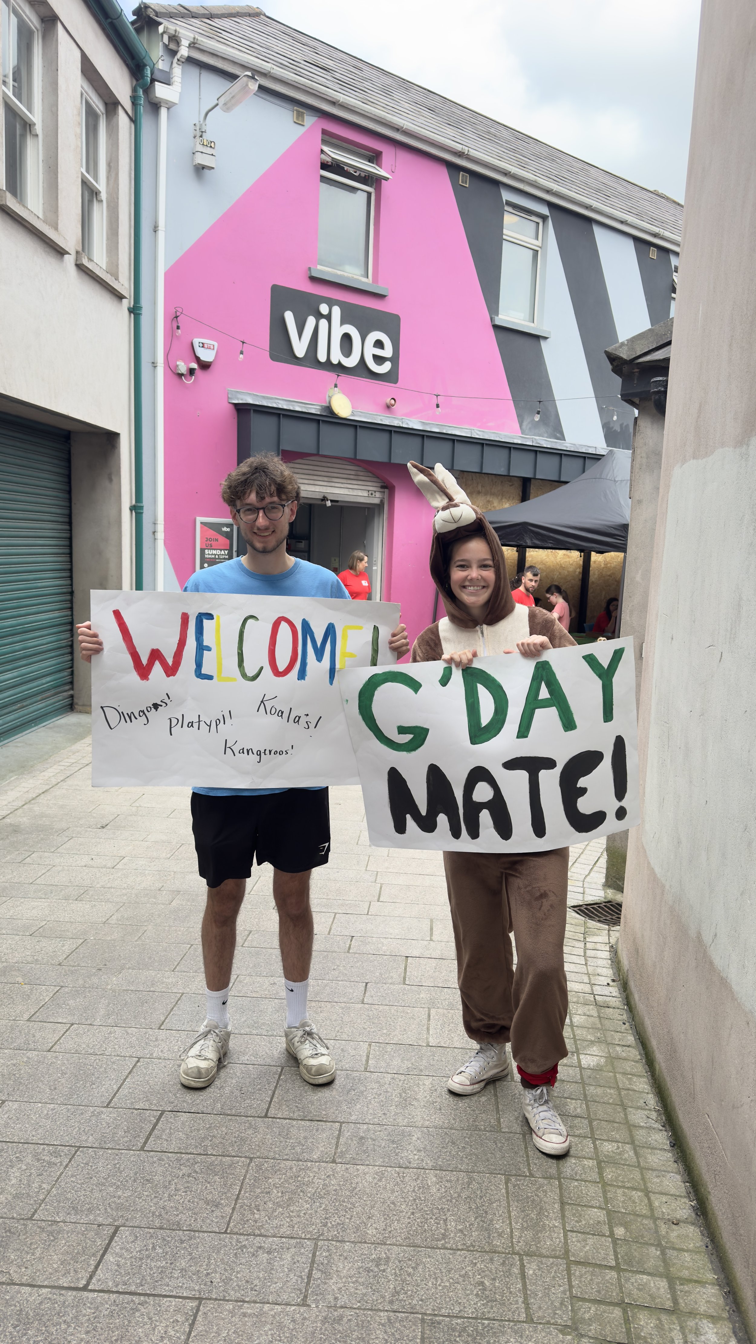Two young people, a man and a woman, are holding colorful signs in a street in front of a pink and black building with a 'vibe' sign. The man is smiling, wearing a blue shirt, black shorts, white socks, and sneakers. The woman, dressed in a bunny costume, also smiling, is holding a sign that says 'G'DAY MATE!' and wearing beige pants and sneakers.